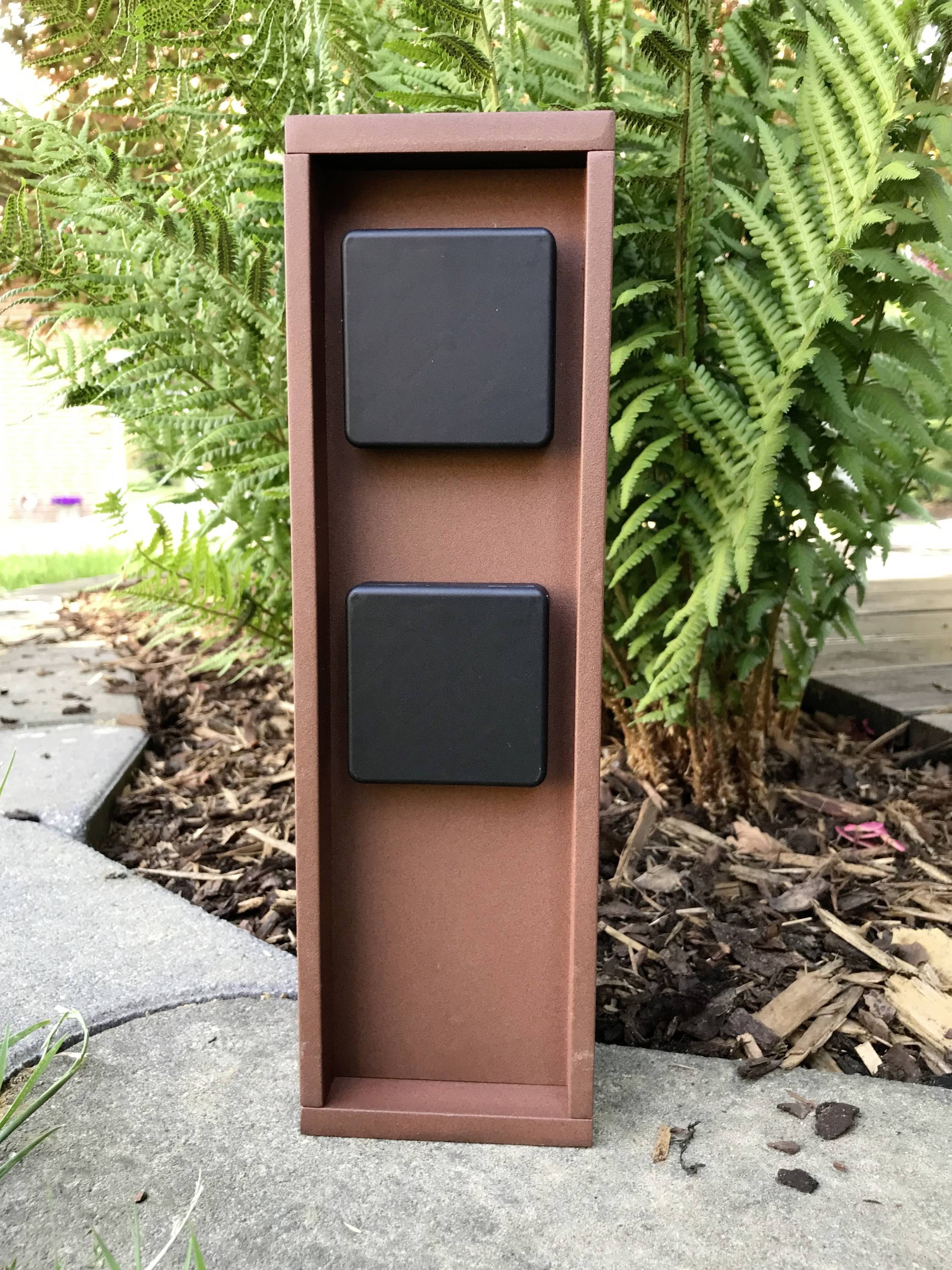 Een rechthoekige, bruine buitenstopcontact met twee zwarte afdekdeksels staat op een bestrate weg voor groene varens in de tuin.