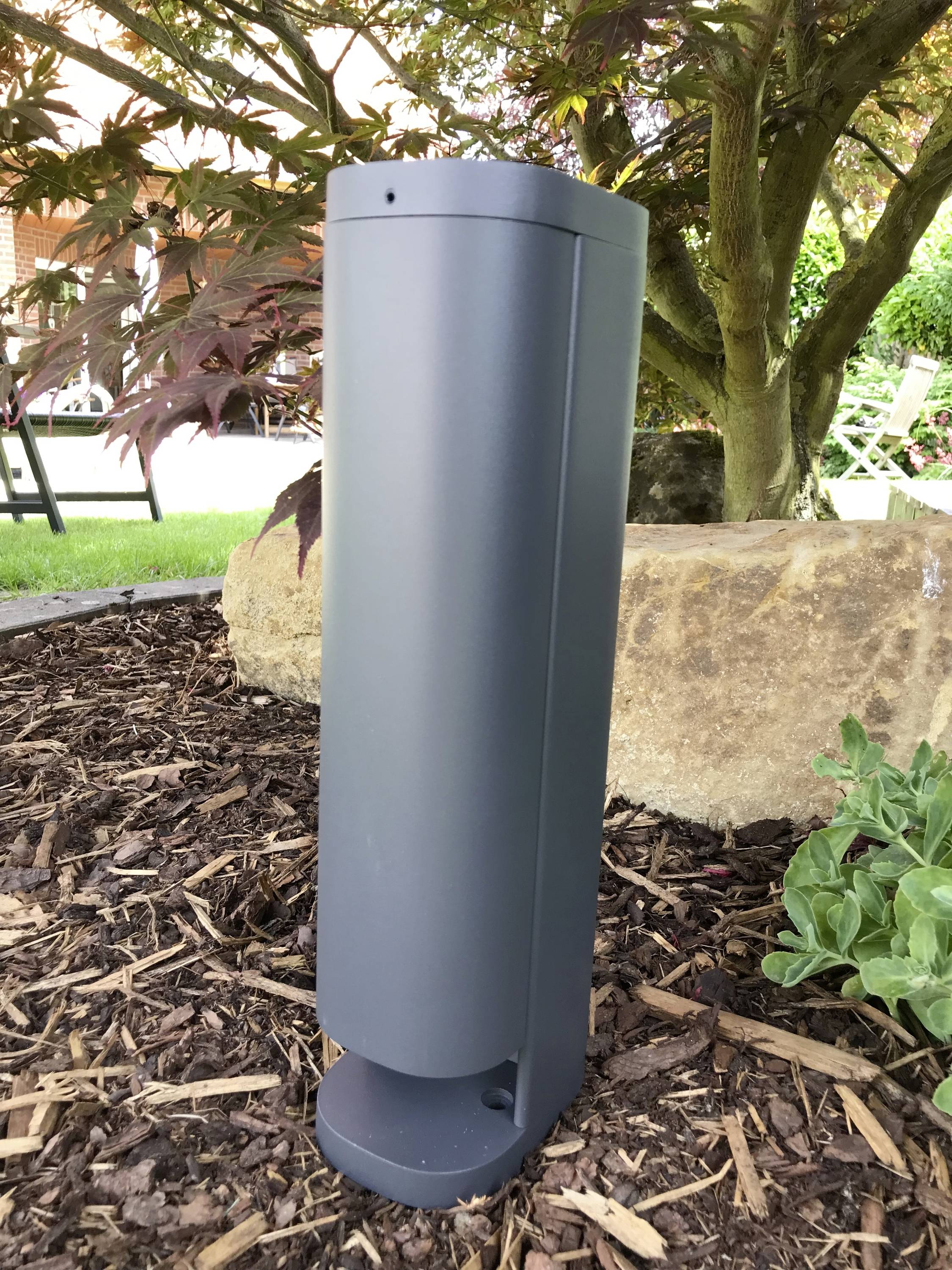 Een grijze metalen lamp staat op bruine mulch naast een steen, omgeven door groene planten en een boom in de tuin.