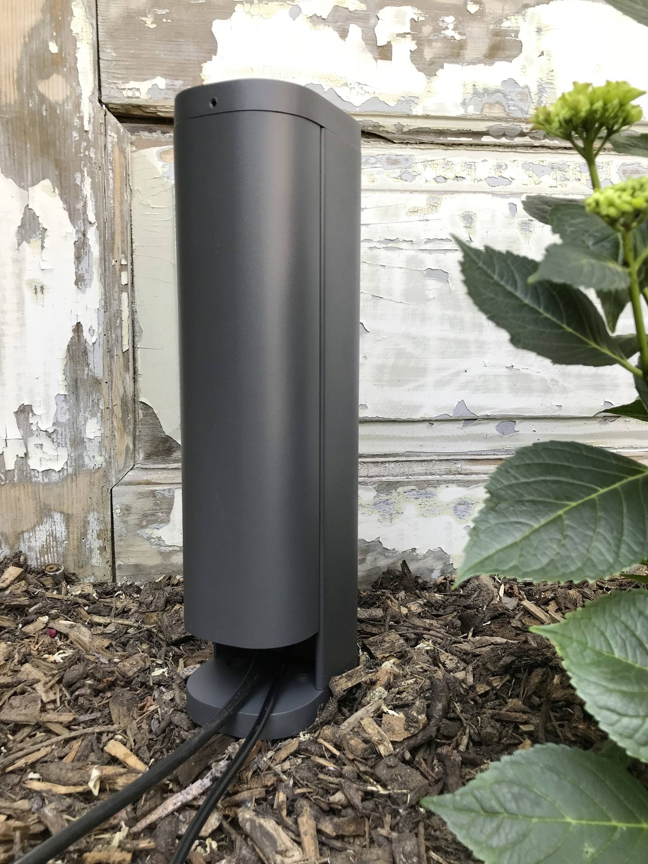 Een zwart, verticaal apparaat staat buiten op de grond naast een bloeiende plant voor een verweerd houten achtergrond.