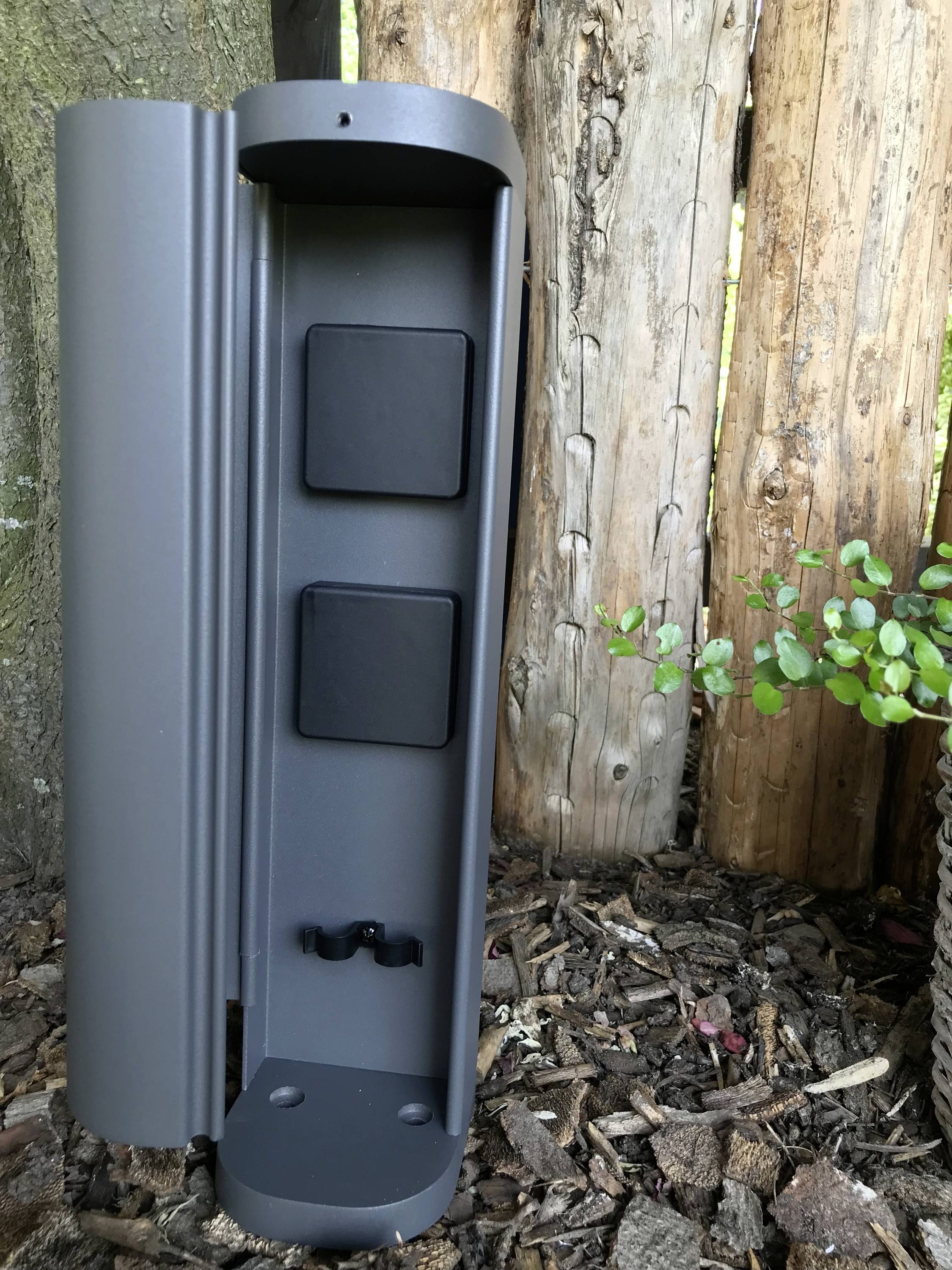 Grijs, open behuizing met twee zwarte vierkanten aan de binnenzijde, gemonteerd op hout in de tuin. Ernaast planten en boomschors.