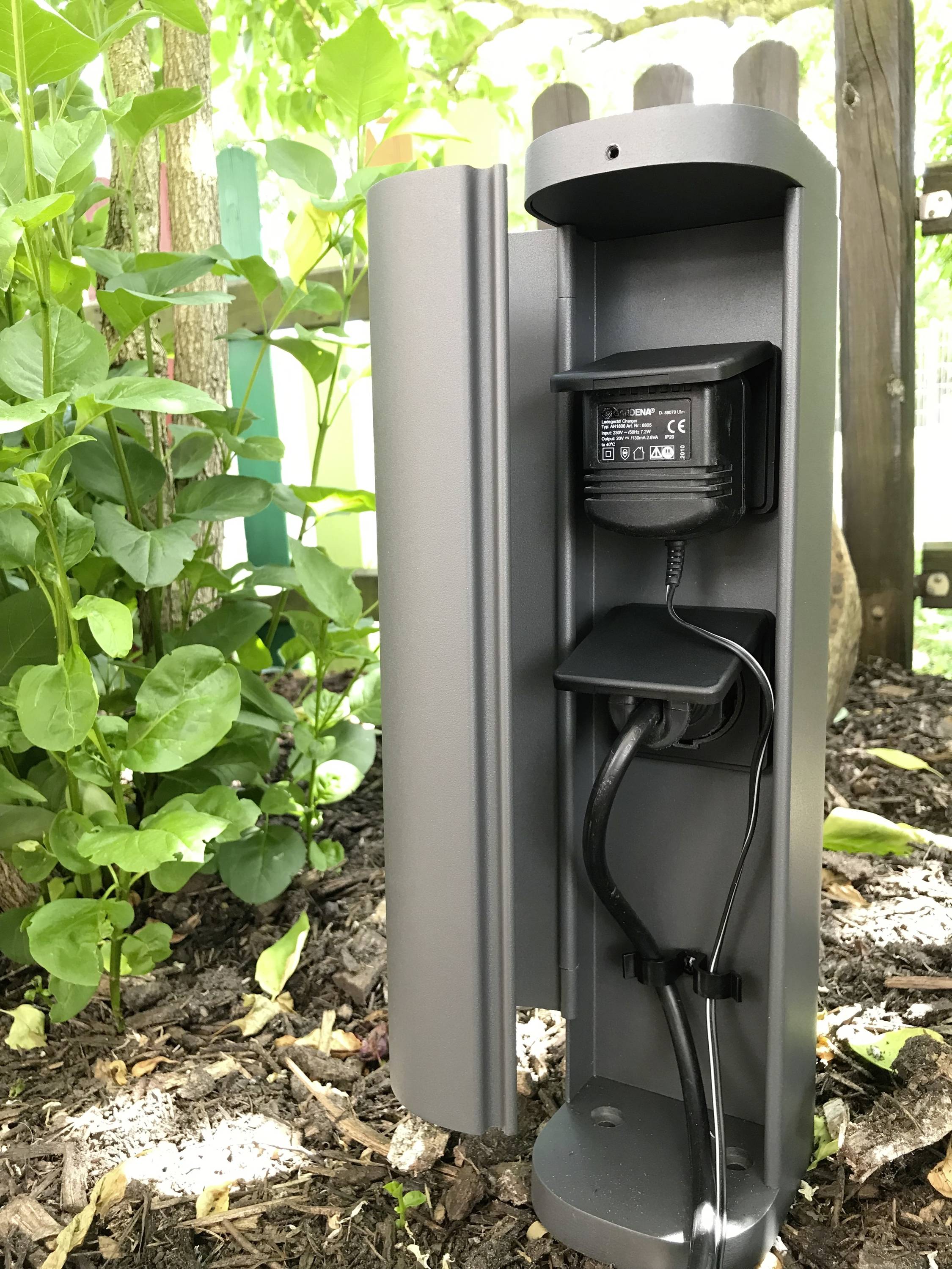 Stopcontact buiten, beschermd in een grijze behuizing, naast planten en aarde, toont kabelaansluitingen. Ideaal voor tuinwerkzaamheden.