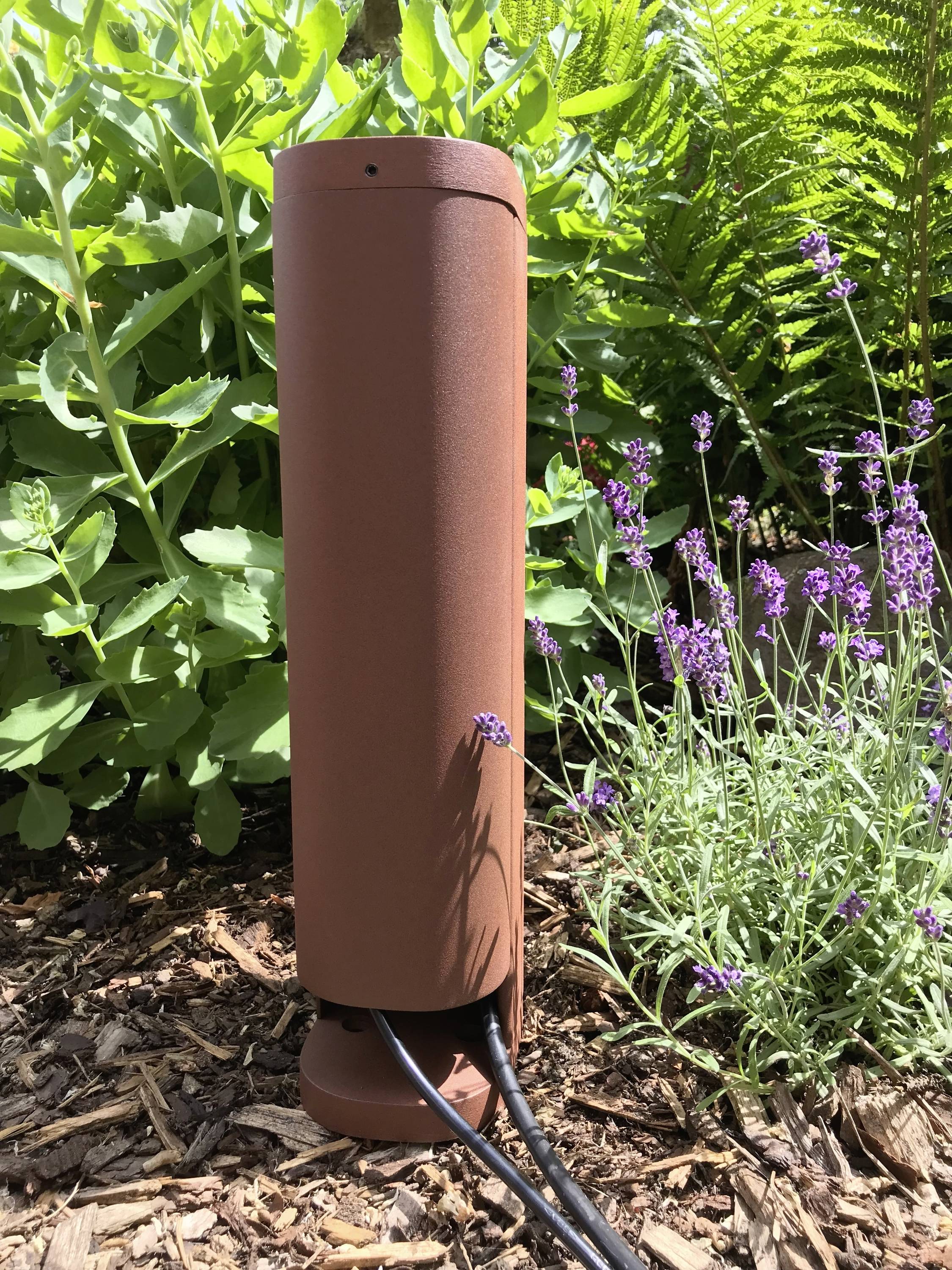 Een bruine, cilindrische tuinlamp staat in een bloembed met lavendel en groene planten. Kabels lopen van de lamp vandaan.