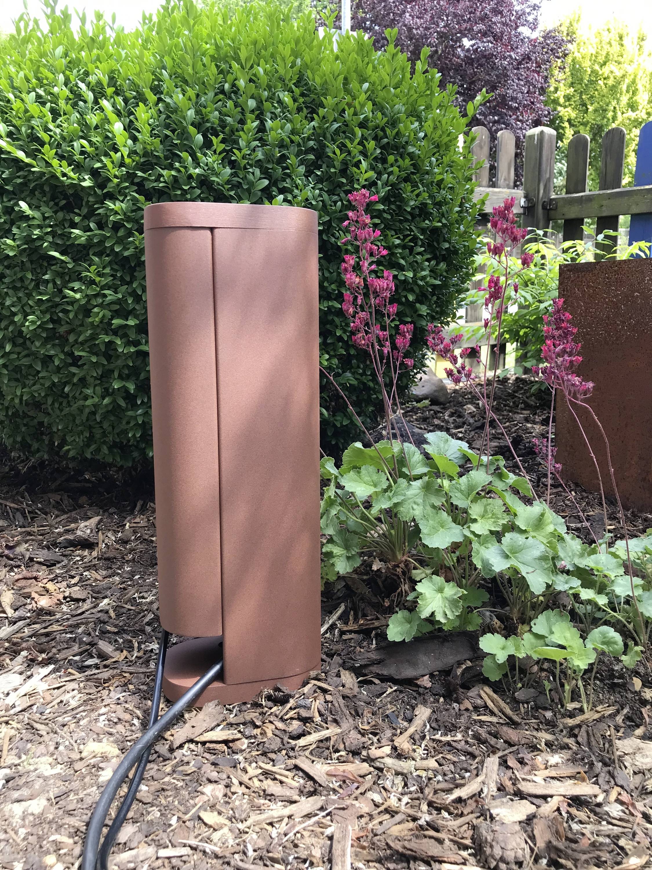 Een bronzen metalen fontein in een tuin, omringd door groene en rode planten, op een mulchondergrond, met een kabel aangesloten.
