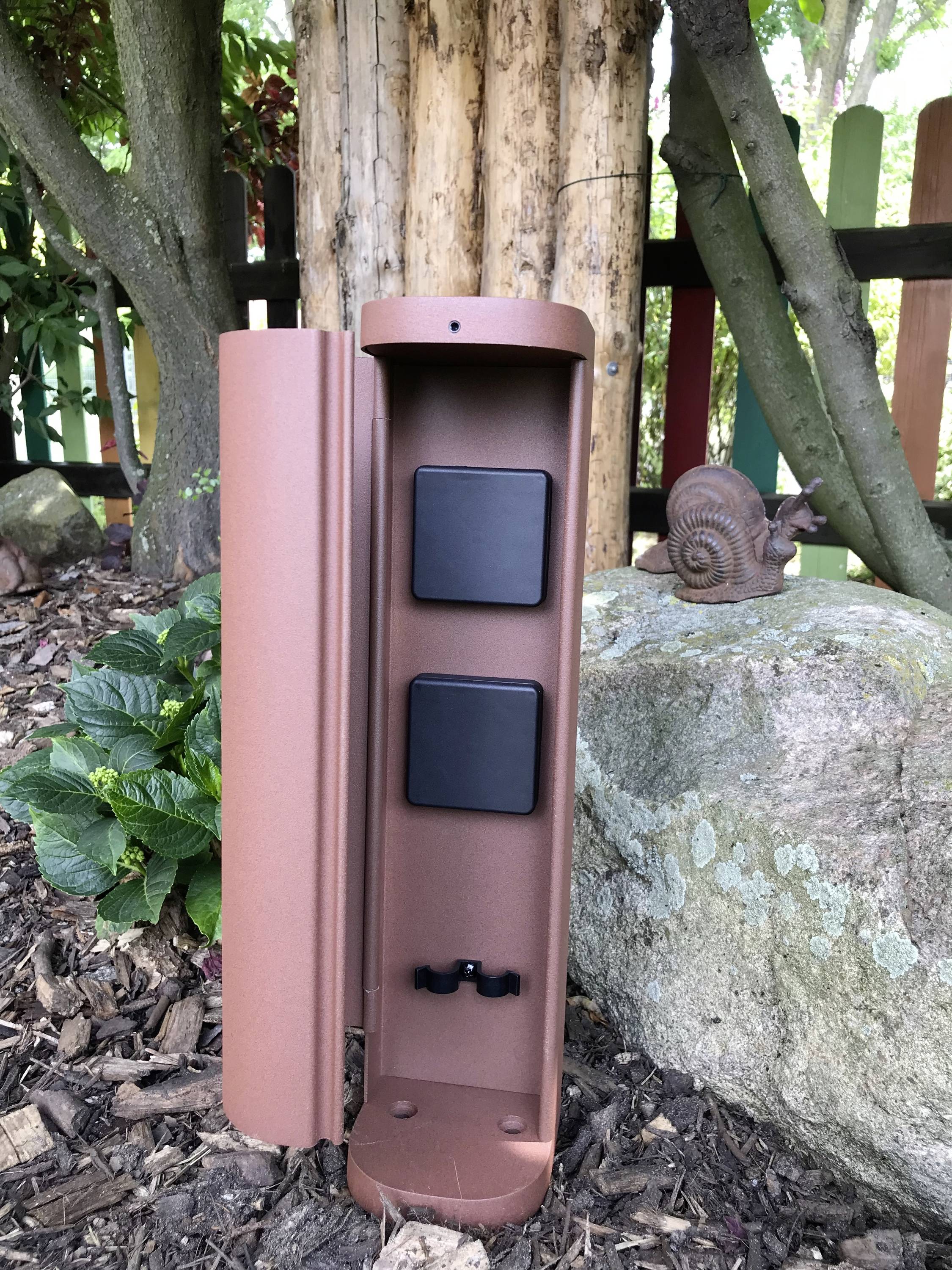 Een bruine metalen doos met twee zwarte schakelaars, gemonteerd naast een rots en een slak in de tuin, omringd door planten.