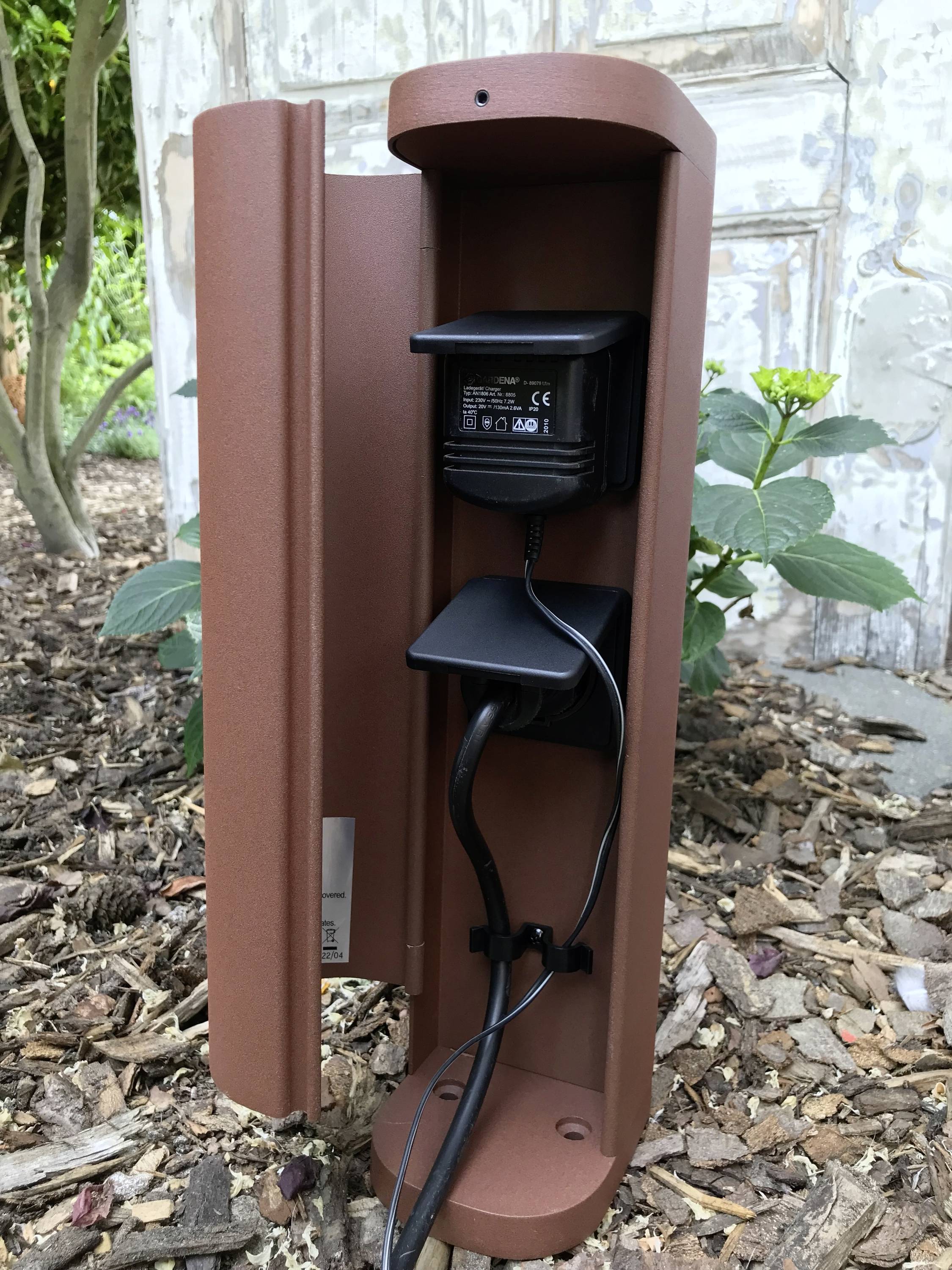 Een bruine buitenstopcontact-zuil met aangesloten stekker staat op de grond voor een houten wand, omringd door planten.