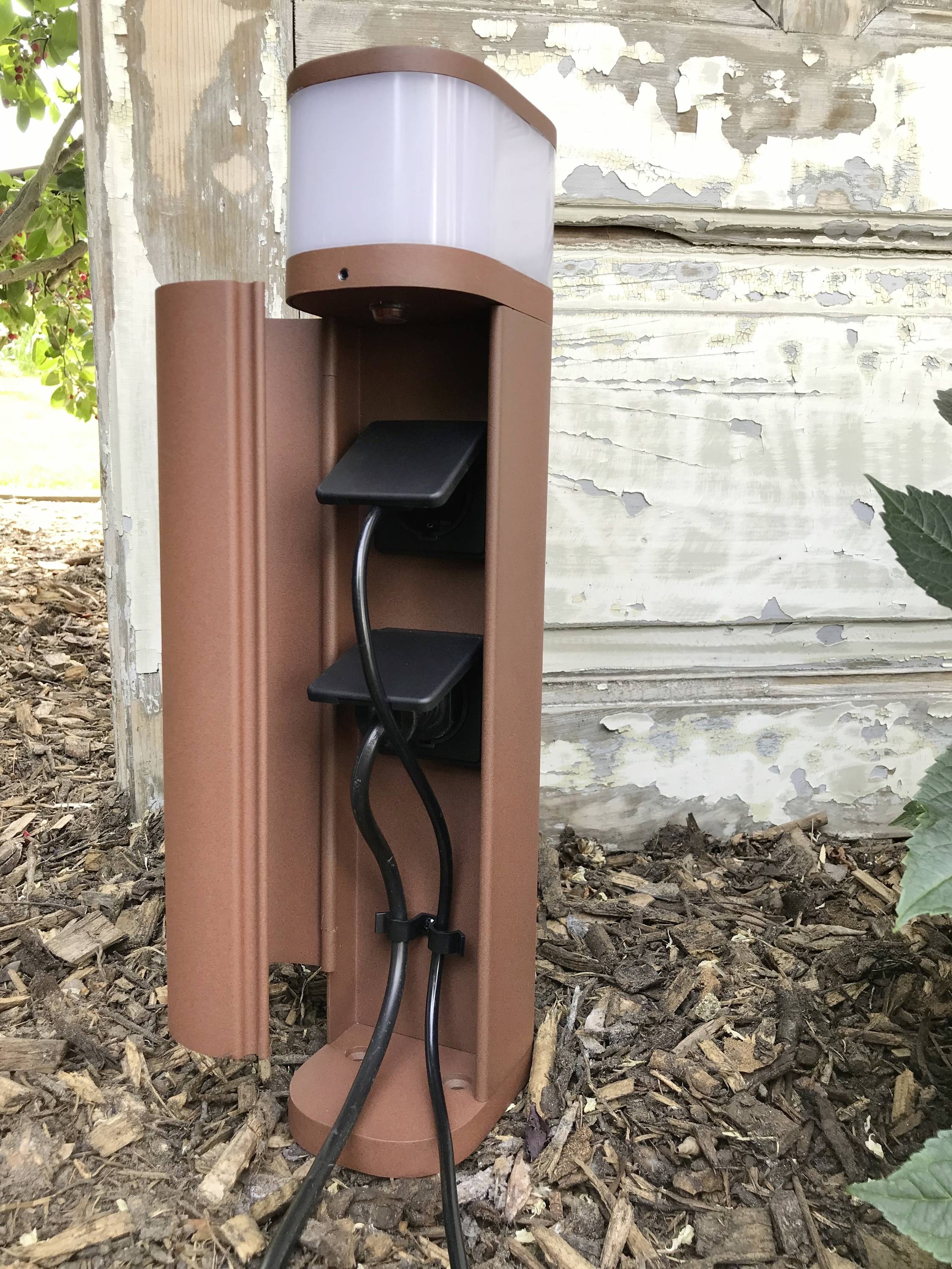 Een bruine contactdoos-zuil met twee zwarte stekkers staat op mulch voor een oude houten wand.