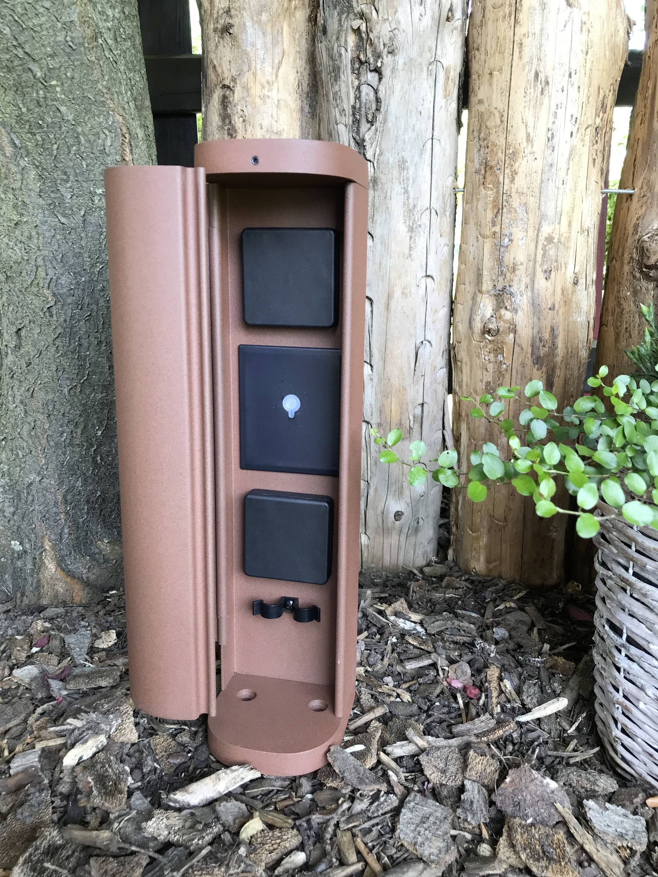 Metalen buitenstroom-aansluitkast met twee stopcontacten en een verlichting, geplaatst op houtsnippers naast een boom en een plant.