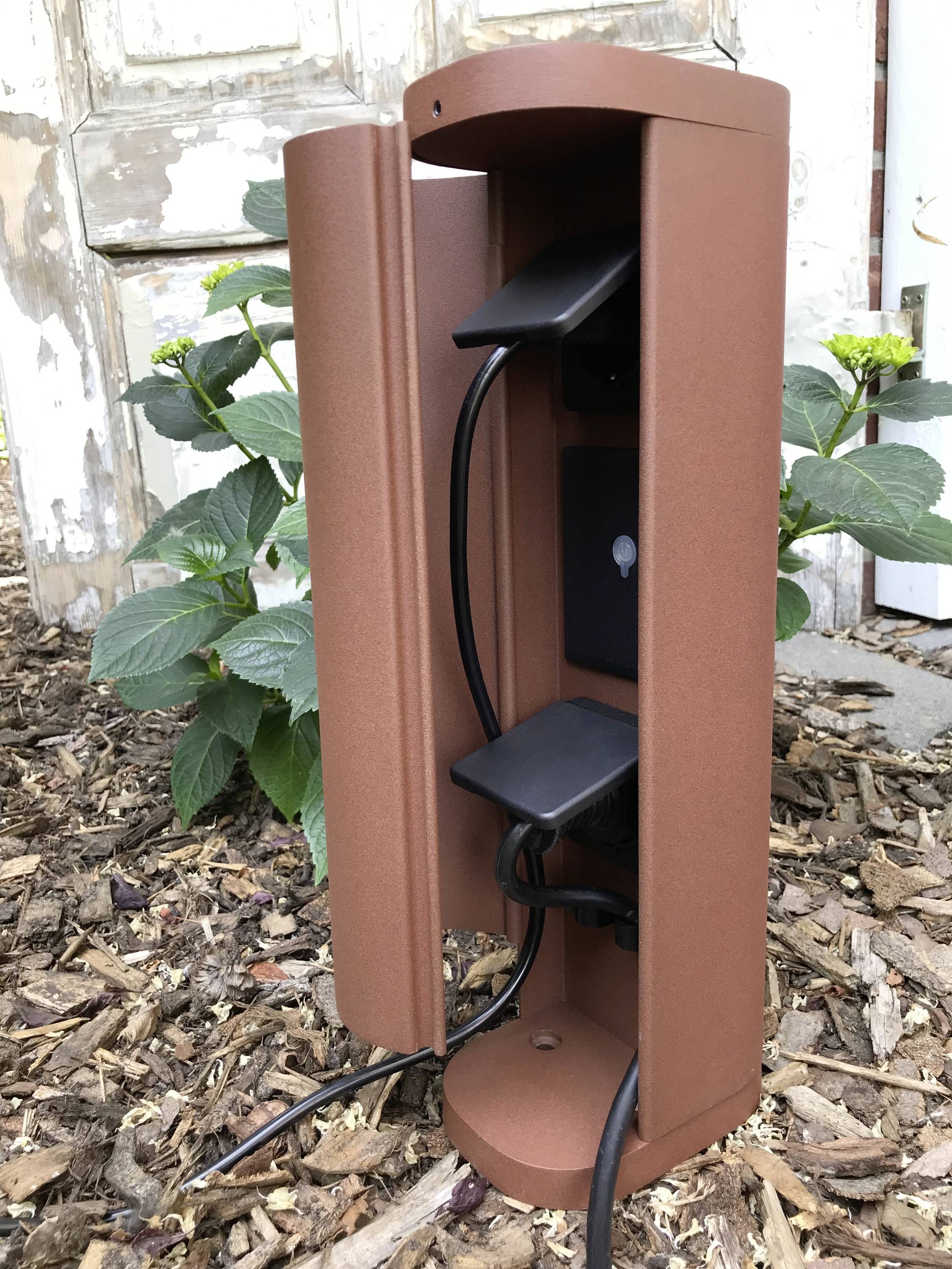 Bruine beschermbox met open kabelgeleiding, geplaatst op mulch naast planten en een houten deur. Ideaal voor tuininstallaties.