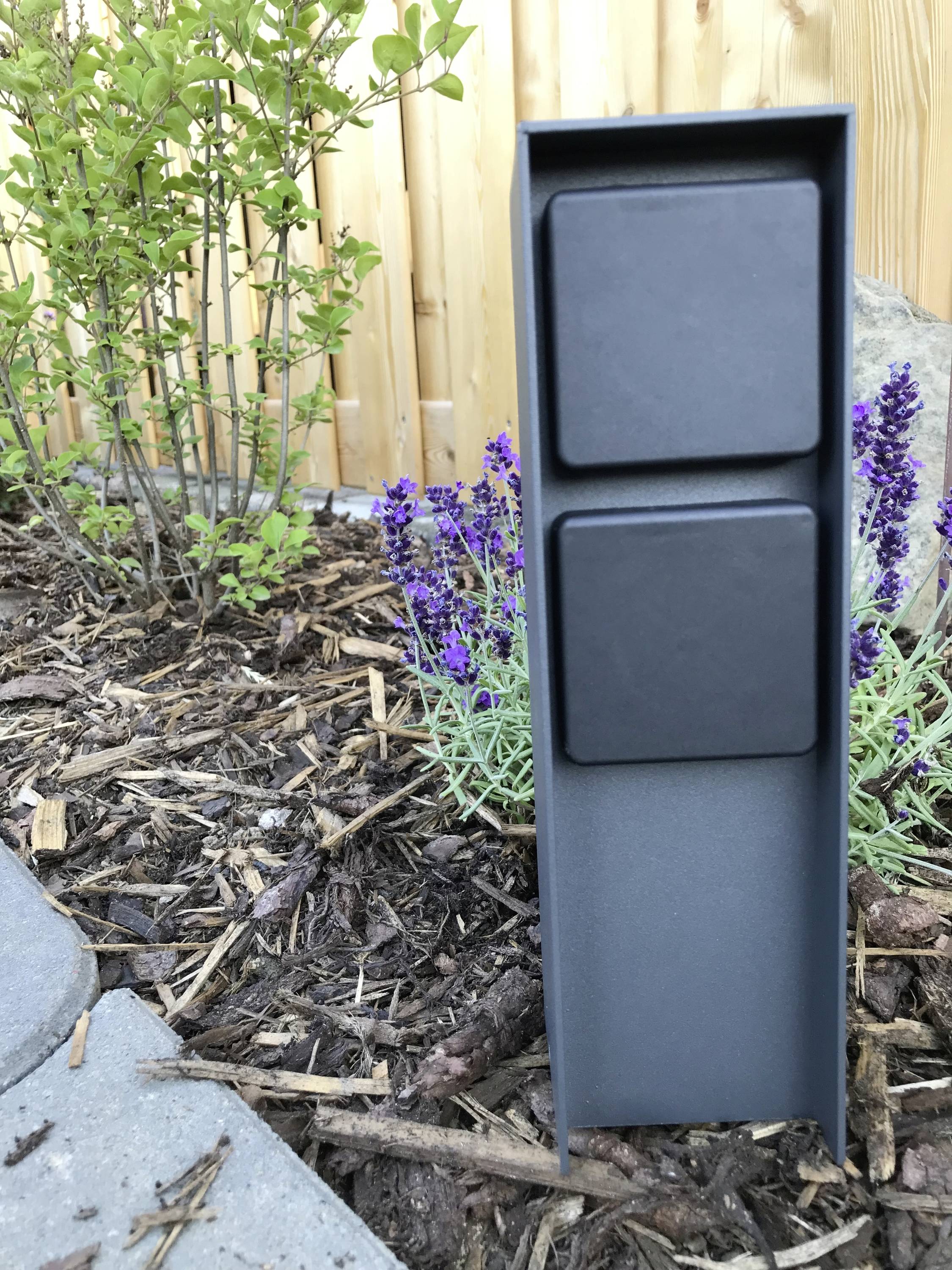 Een grijze buitenstopcontact staat in een bloembed naast lavendel en een houten muur, omgeven door mulch en planten.