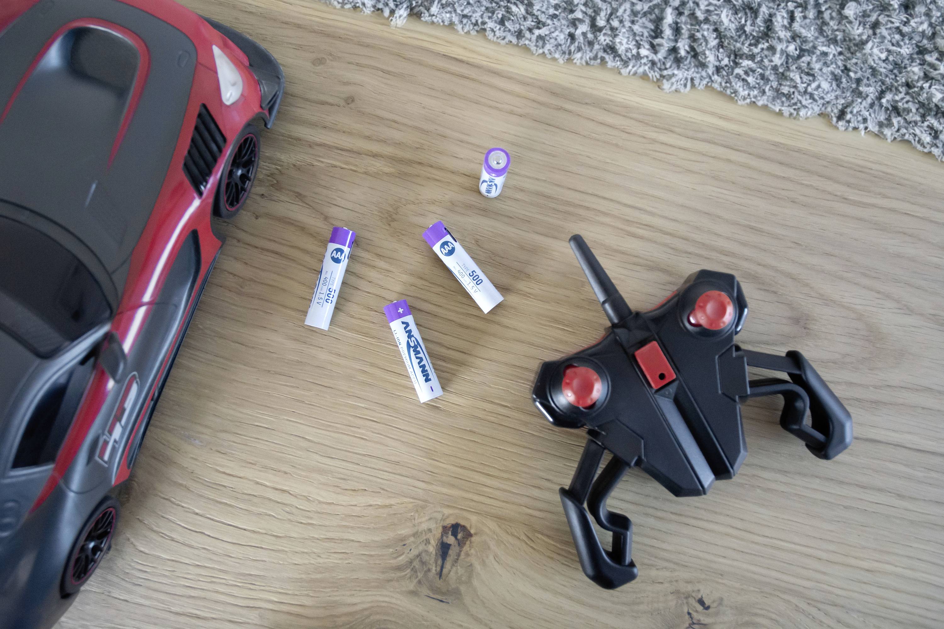 Een rood speelgoedautootje en een zwarte afstandsbediening liggen op een houten vloer. Vier AA-batterijen zijn ernaast verspreid.