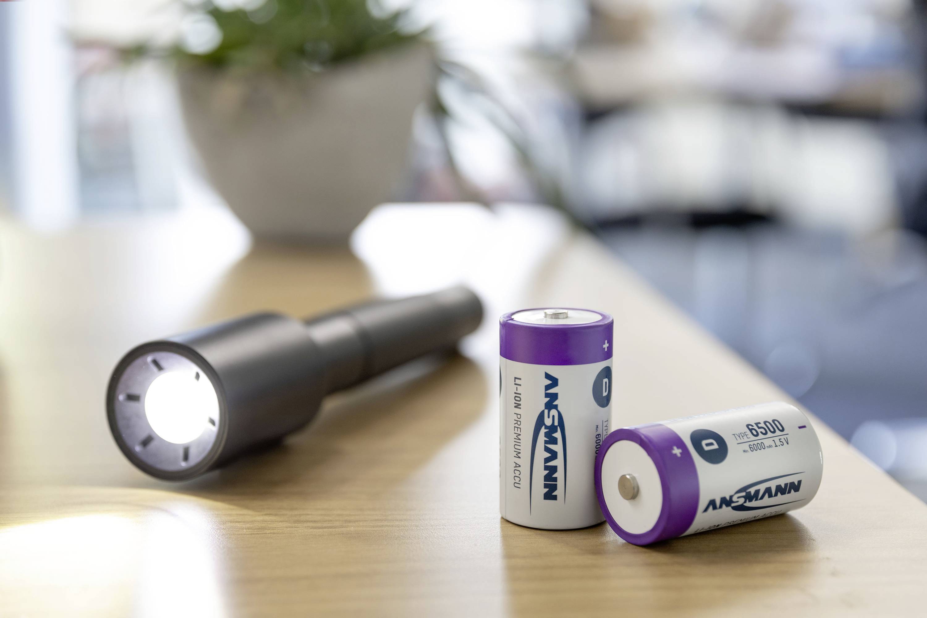 Zaklamp op tafel met twee batterijen ernaast, klaar voor gebruik. Op de achtergrond een plant onscherp zichtbaar.