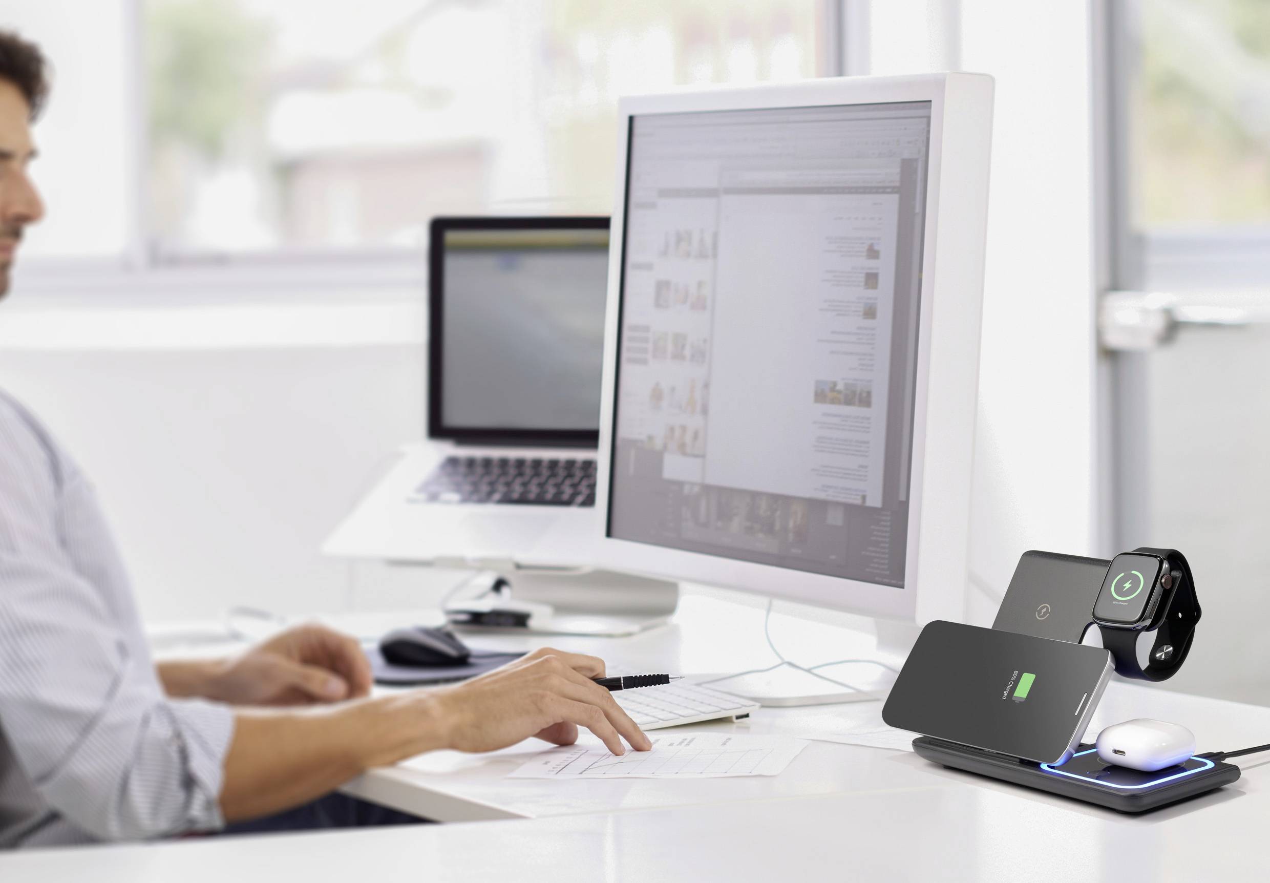 Een man werkt op de computer in een modern kantoor. Op de voorgrond staat een oplaadstation met een smartphone en een smartwatch.