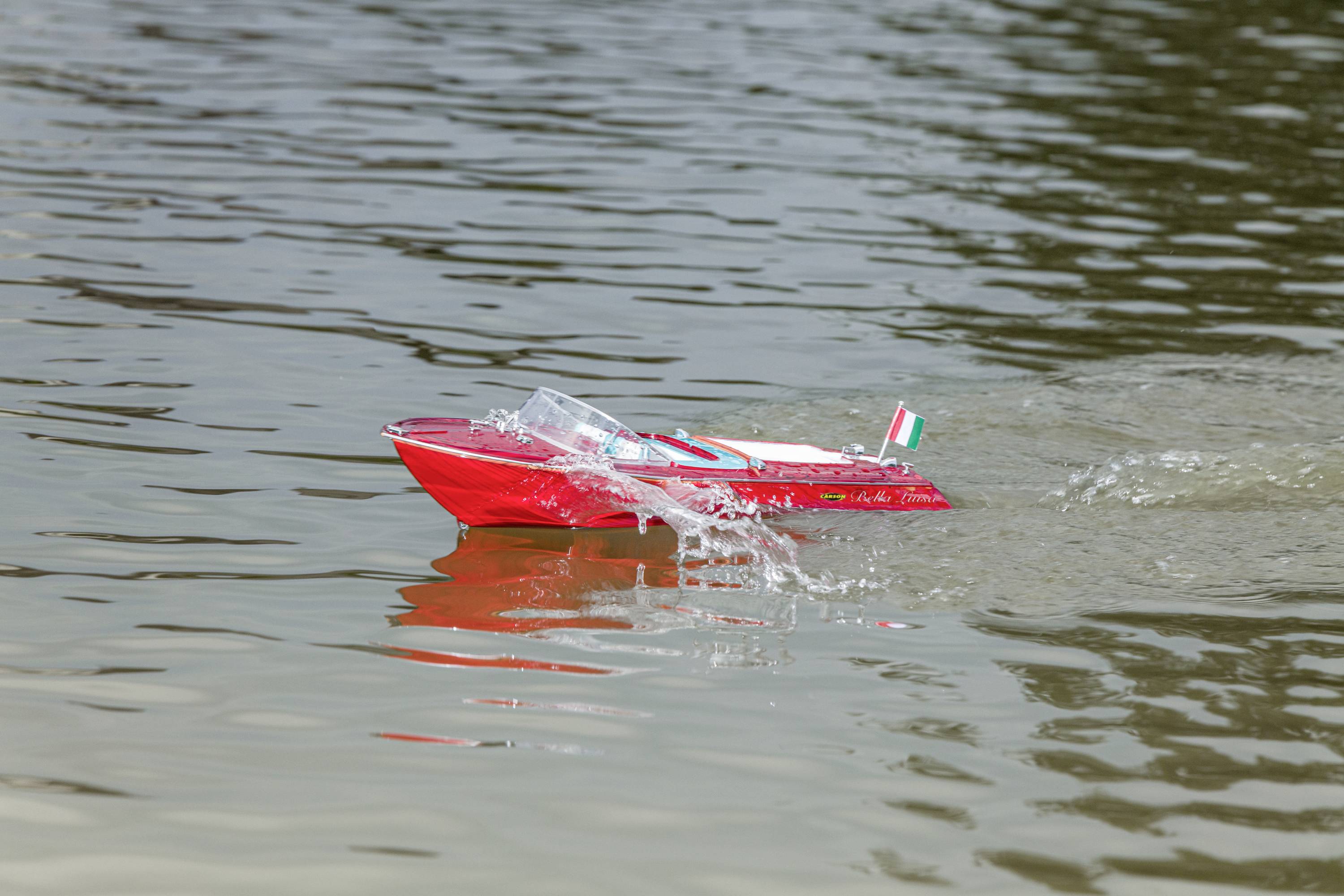 Een kleine rode modelboot met een Italiaanse vlag vaart over een meer en veroorzaakt lichte golven.