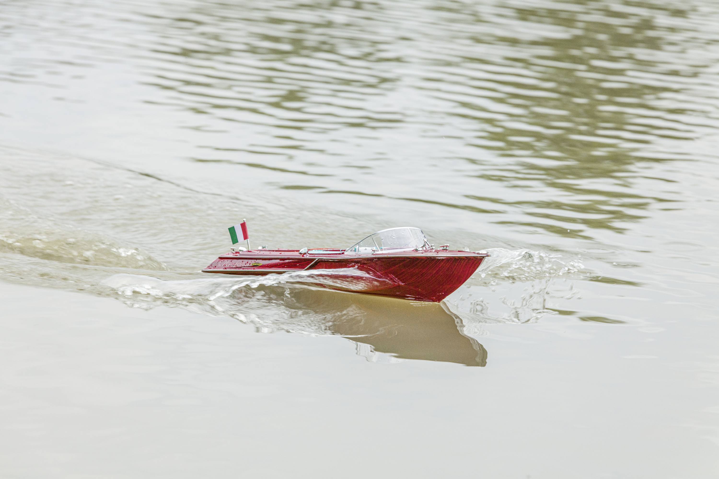 Een rood modelboot met een Italiaanse vlag vaart op een rustig water.
