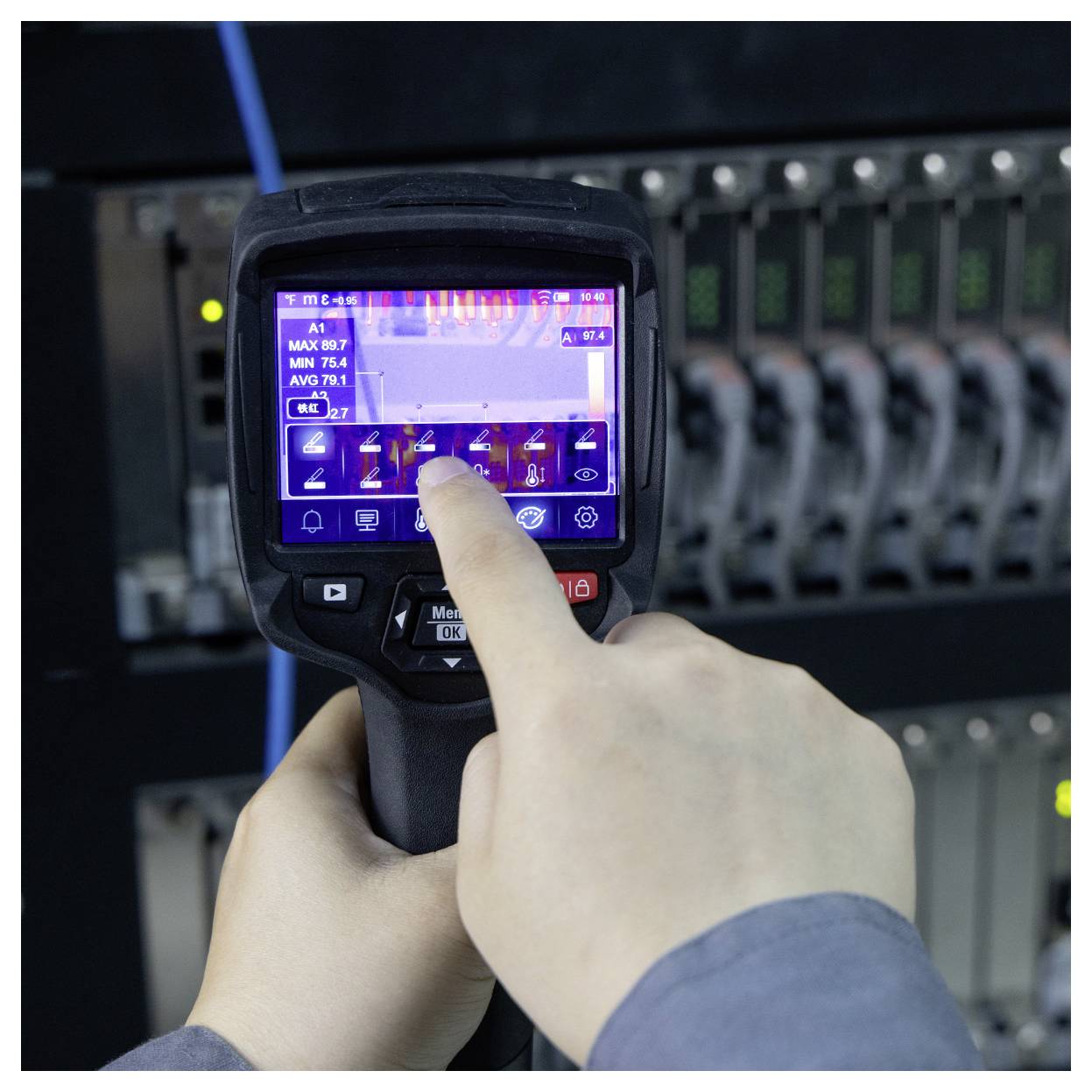 Persoon die een handheld thermische beeldcamera gebruikt om een elektrisch bedieningspaneel te inspecteren, met op het scherm een hittemap en temperatuurmetingen.