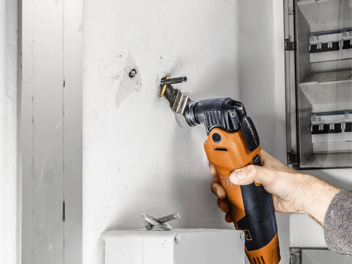 Een hand houdt een oscillerend multifunctioneel gereedschap vast dat een muurhaak doorsnijdt. Op de achtergrond is een open verdeeldoos zichtbaar.