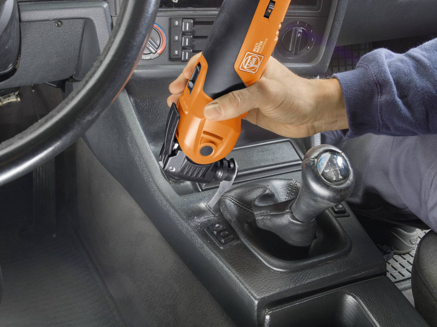 Een persoon reinigt de middenconsole van een auto met een oranje, accugevoed multitool.