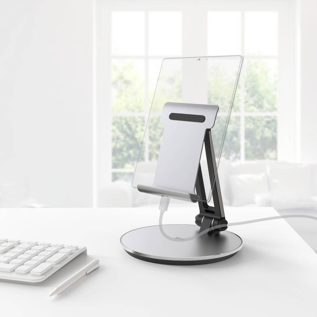 Tablet op een standaard naast een toetsenbord op een bureau, met een raam op de achtergrond.