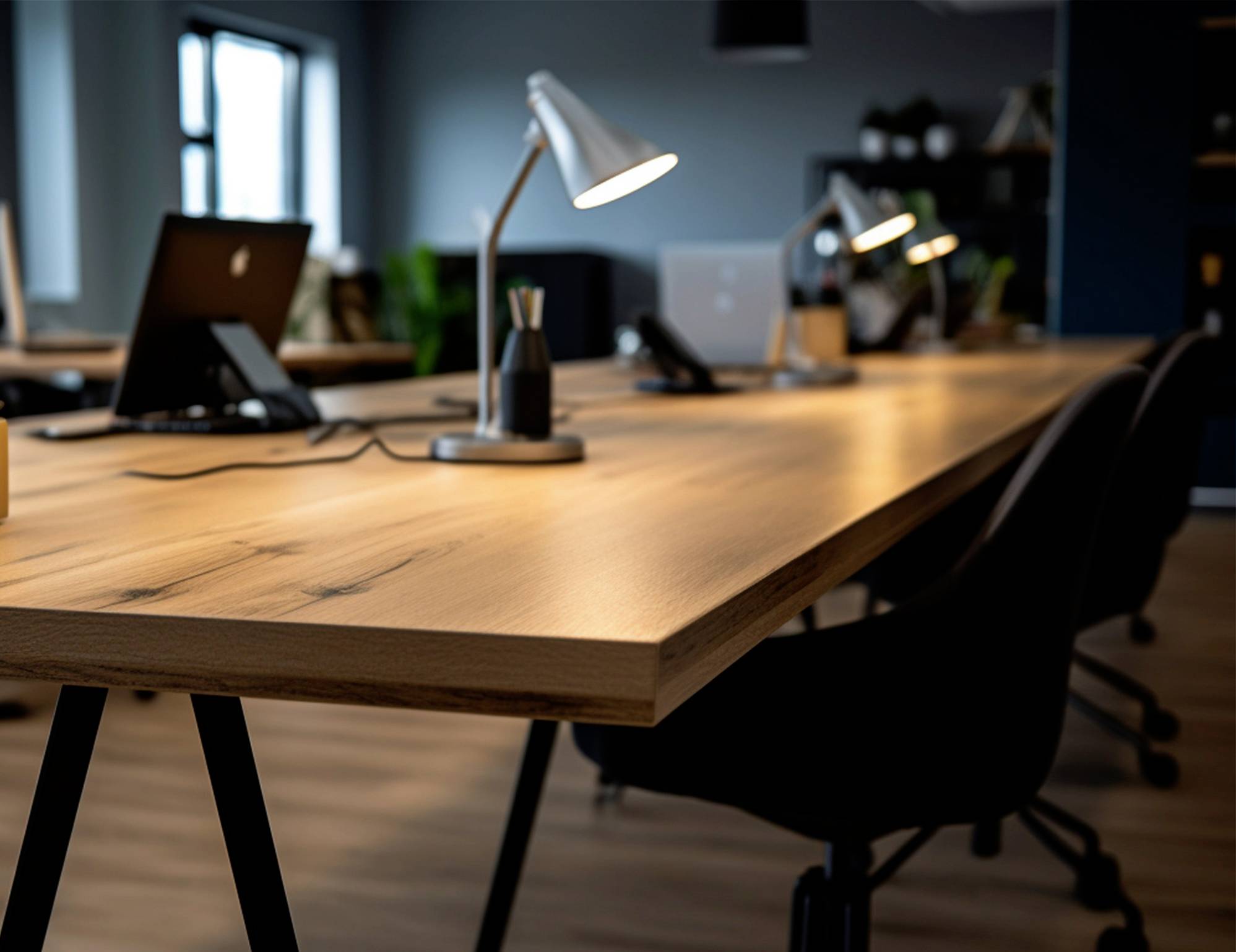 Een moderne, lege kantoorruimte met een lange houten tafel, waarop drie bureaulampen en laptops staan. Op de achtergrond zijn grote ramen.