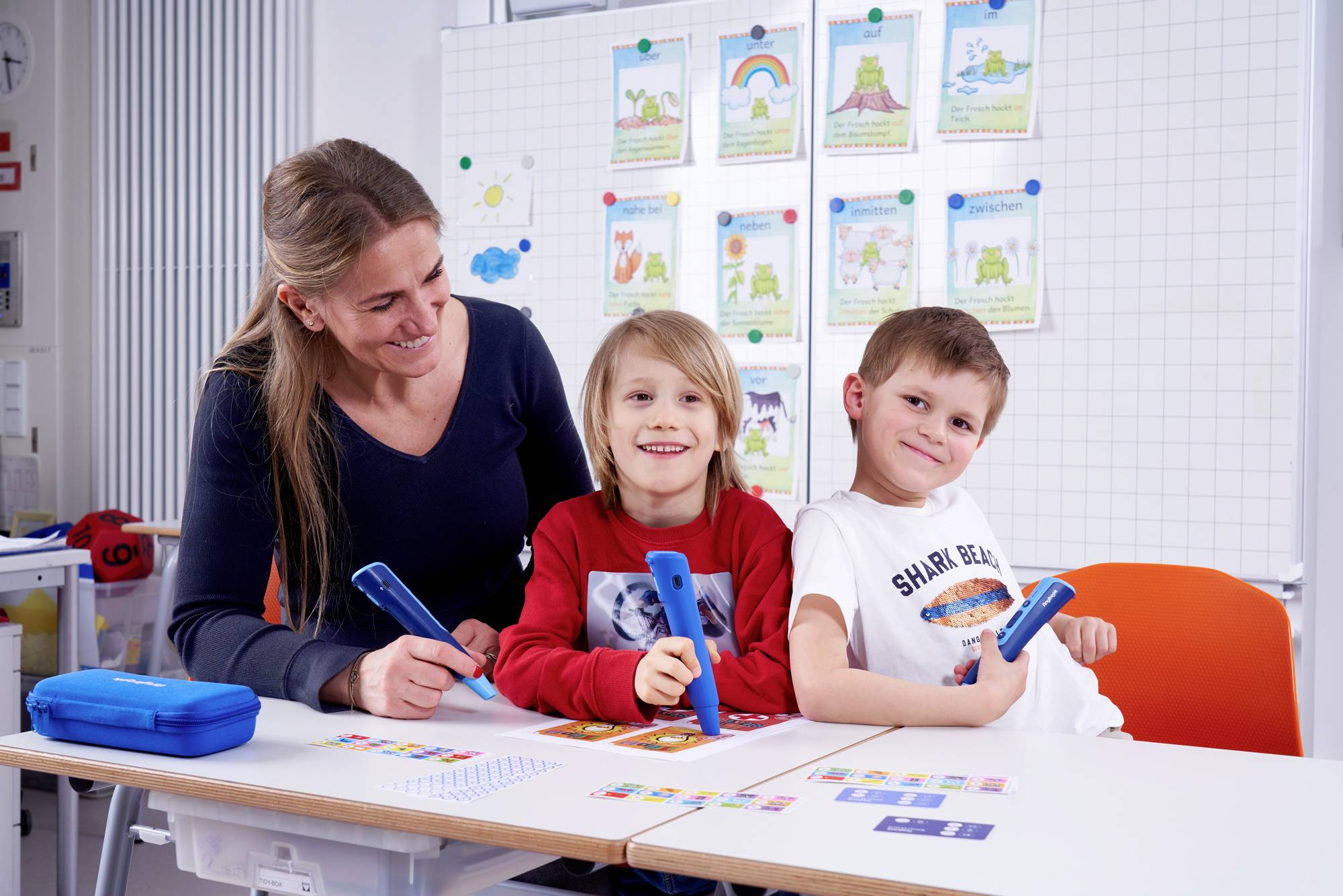 Een lerares helpt twee kinderen met leren met elektronische pennen in een klaslokaal. Op de achtergrond zijn educatieve posters.