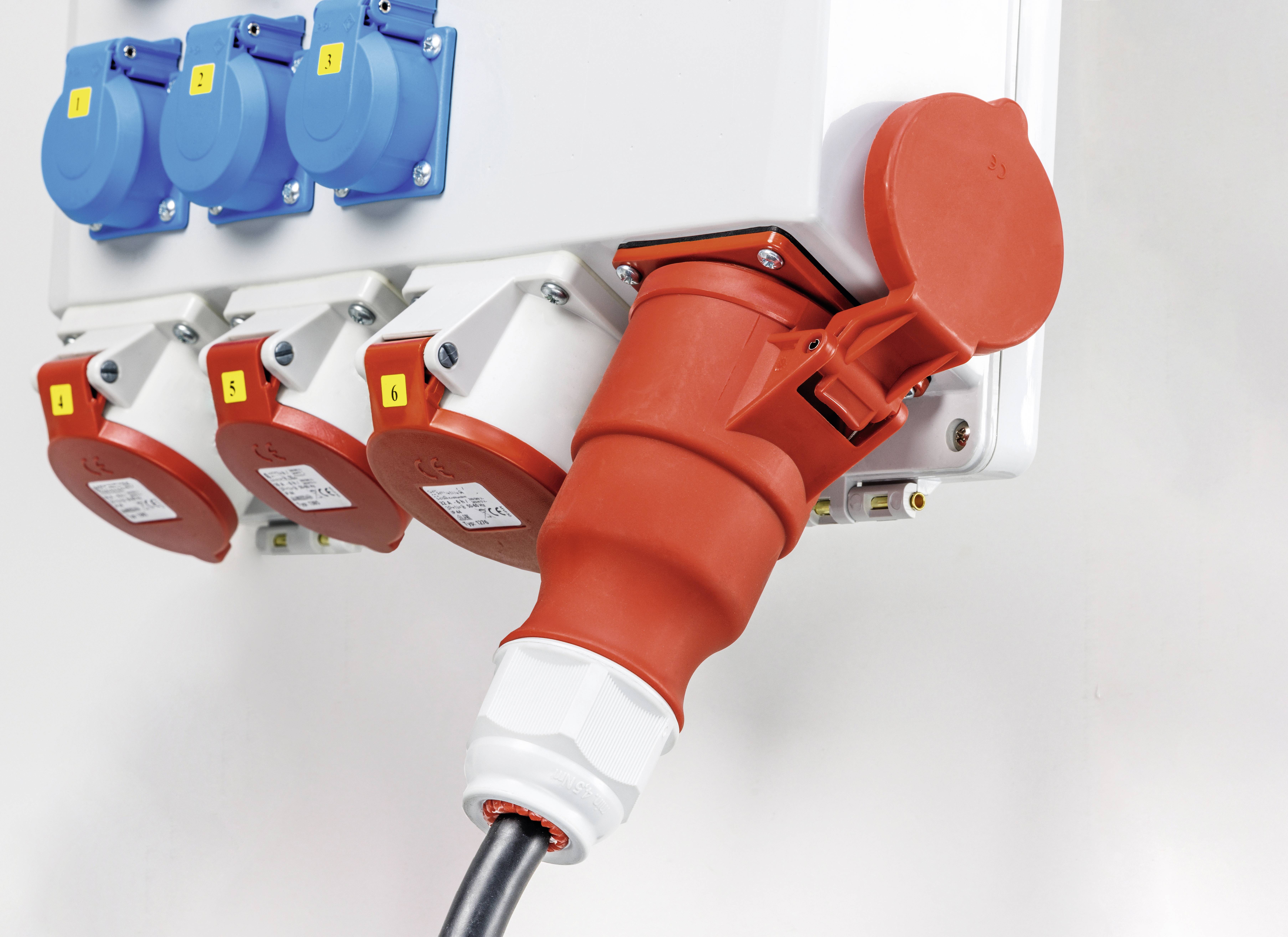 Een rode, industriële elektrische koppeling is gestoken in een witte verdeeldoos. Ernaast bevinden zich blauwe en rode stopcontacten.