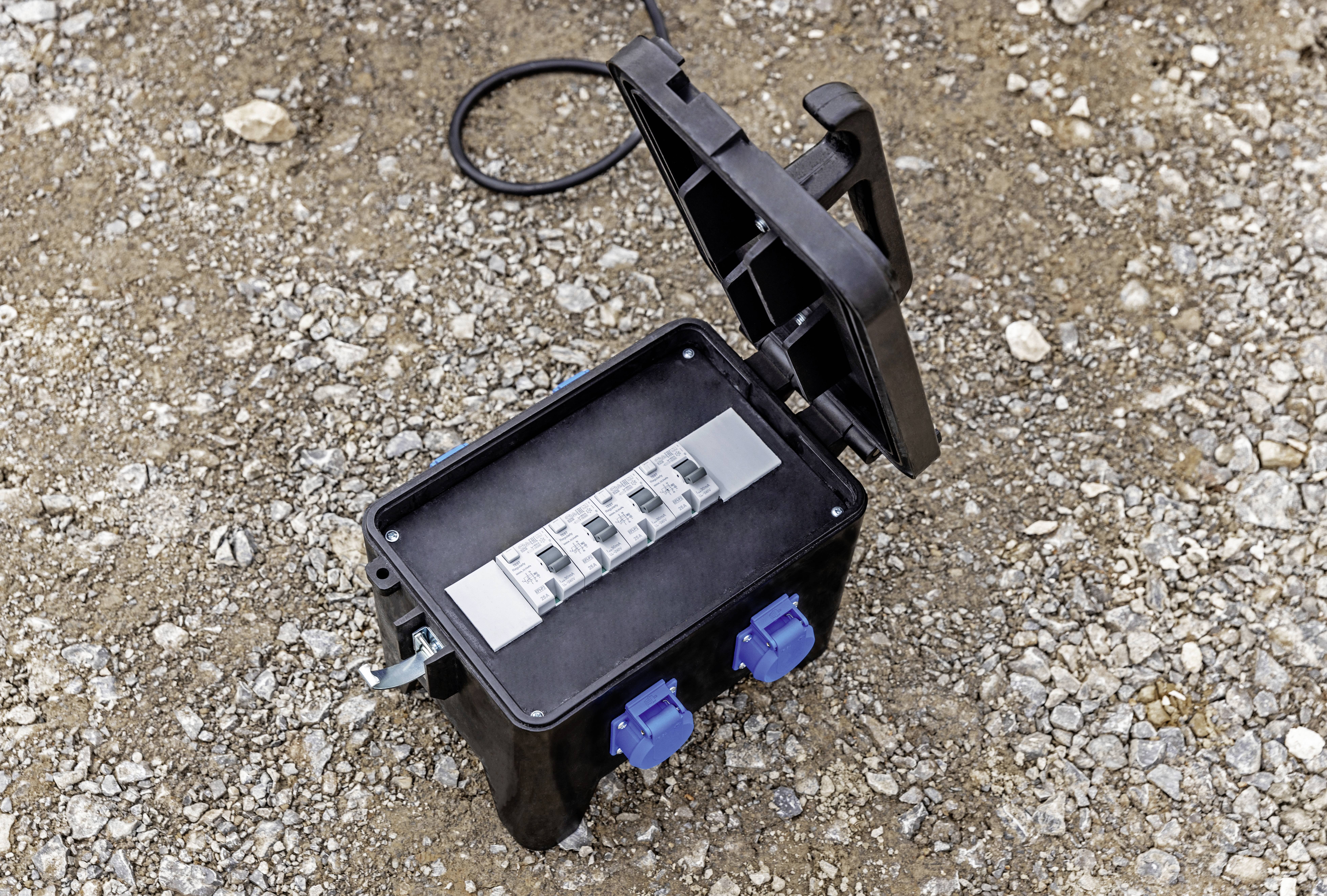 Een open zwarte elektrische aansluitdoos met meerdere kabelaansluitingen en schakelaars ligt op een kiezelige ondergrond.