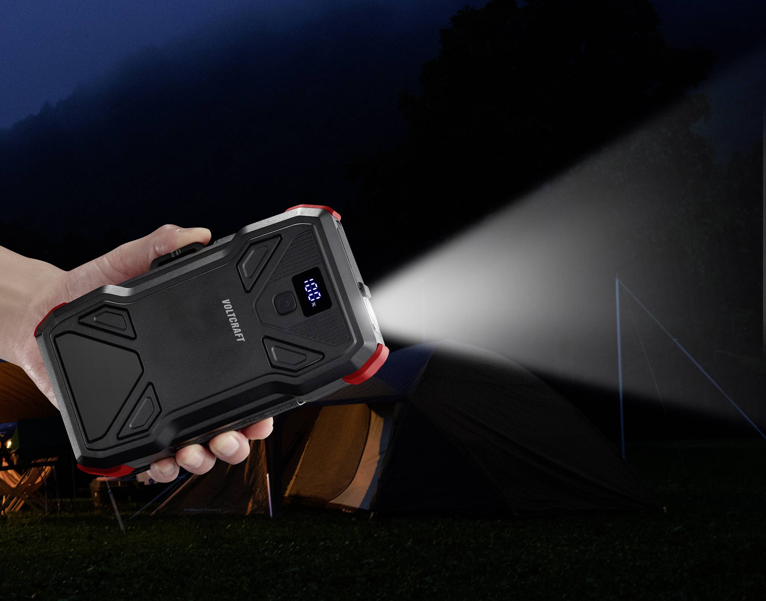 Een hand houdt een draagbare powerbank vast die ook als zaklamp wordt gebruikt. Een tent staat op de achtergrond in het donker.