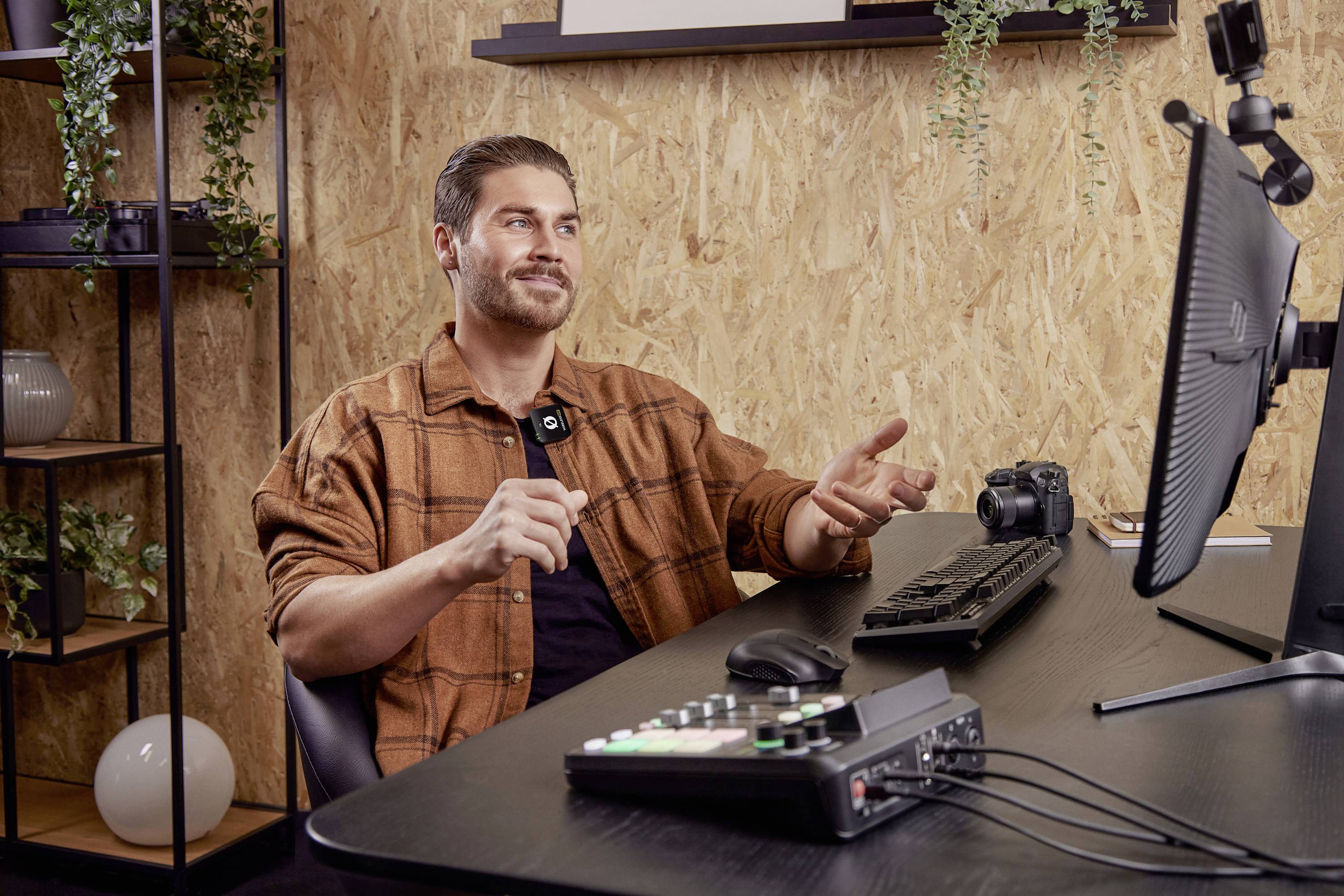 Een man zit aan een bureau met een computer en opnameapparatuur, spreekt in een camera. Kantoorinrichting met houten panelen.