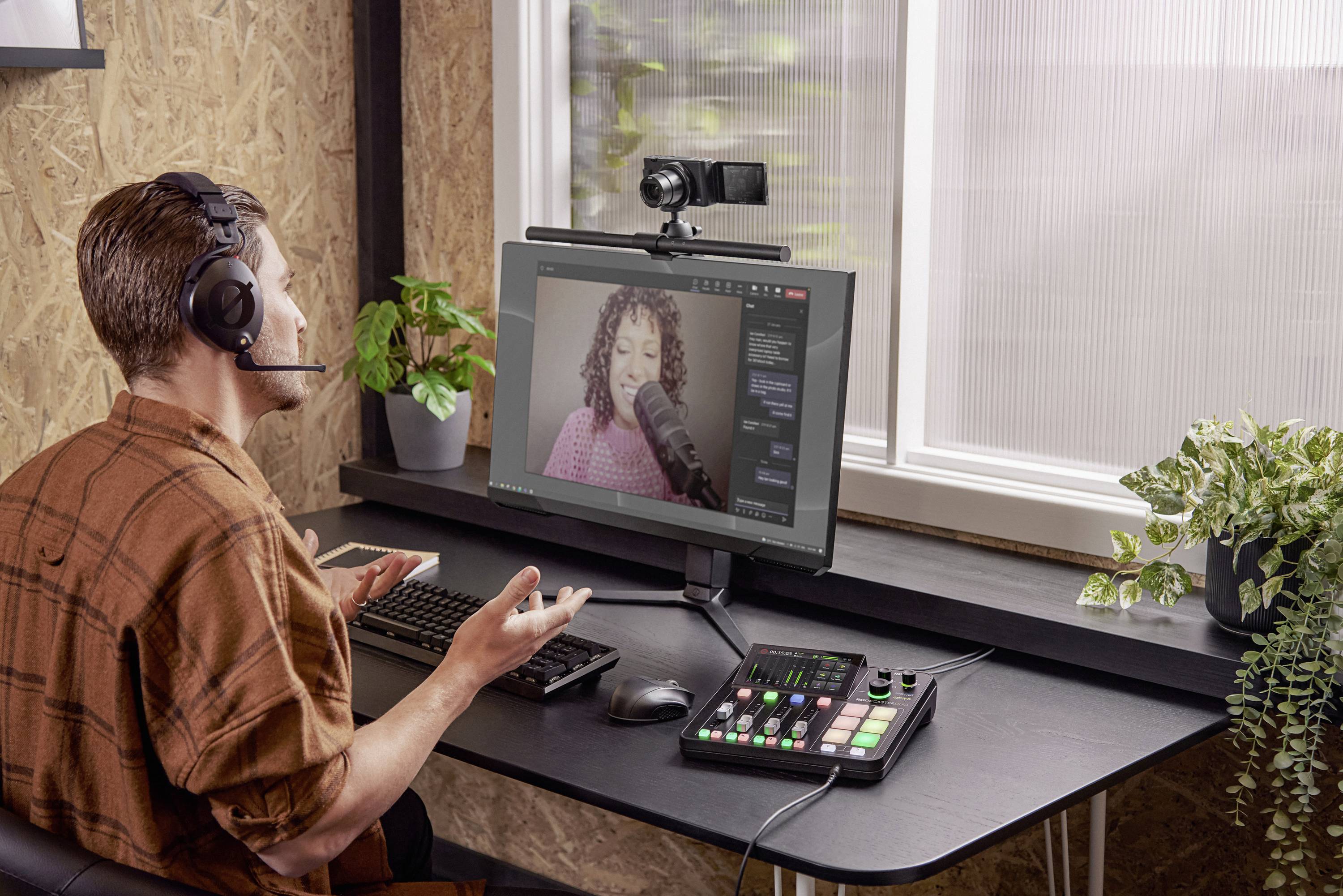 Een man met een headset zit aan een tafel, bedient een computer en een mengpaneel. Op het scherm een vrouw tijdens de opname.