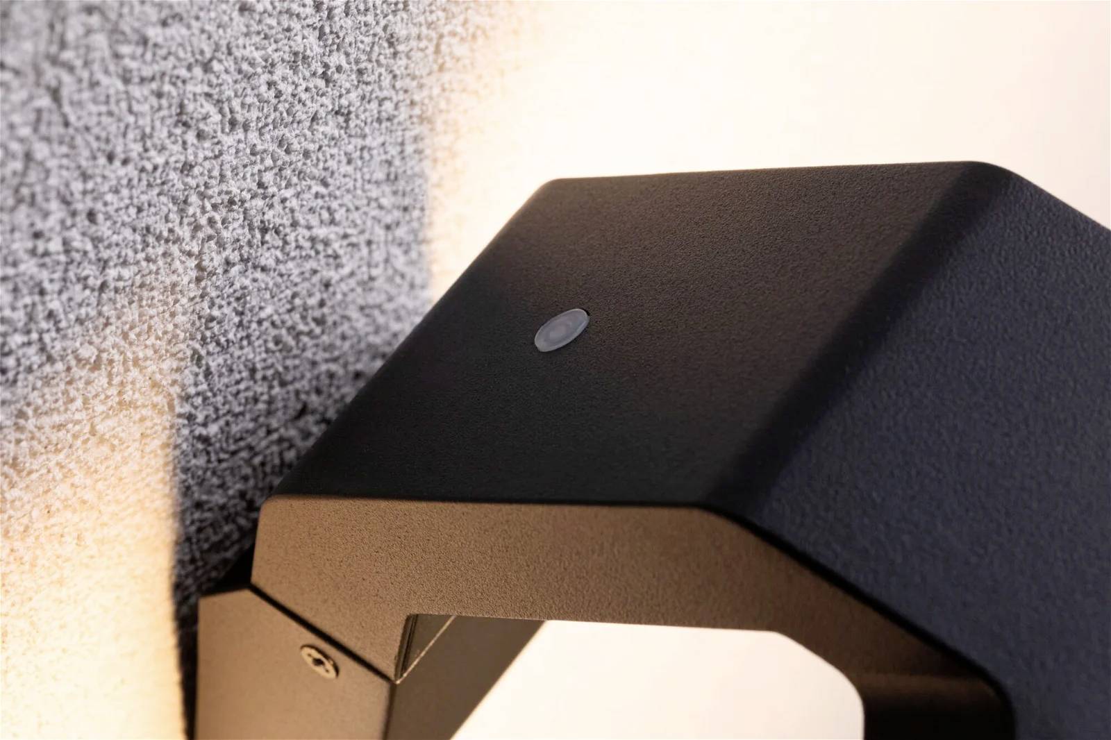 Wandlamp met modern, zwart design steekt uit van een gestructureerde muur. Hij straalt neerwaarts gerichte verlichting uit en creëert een zachte sfeer.