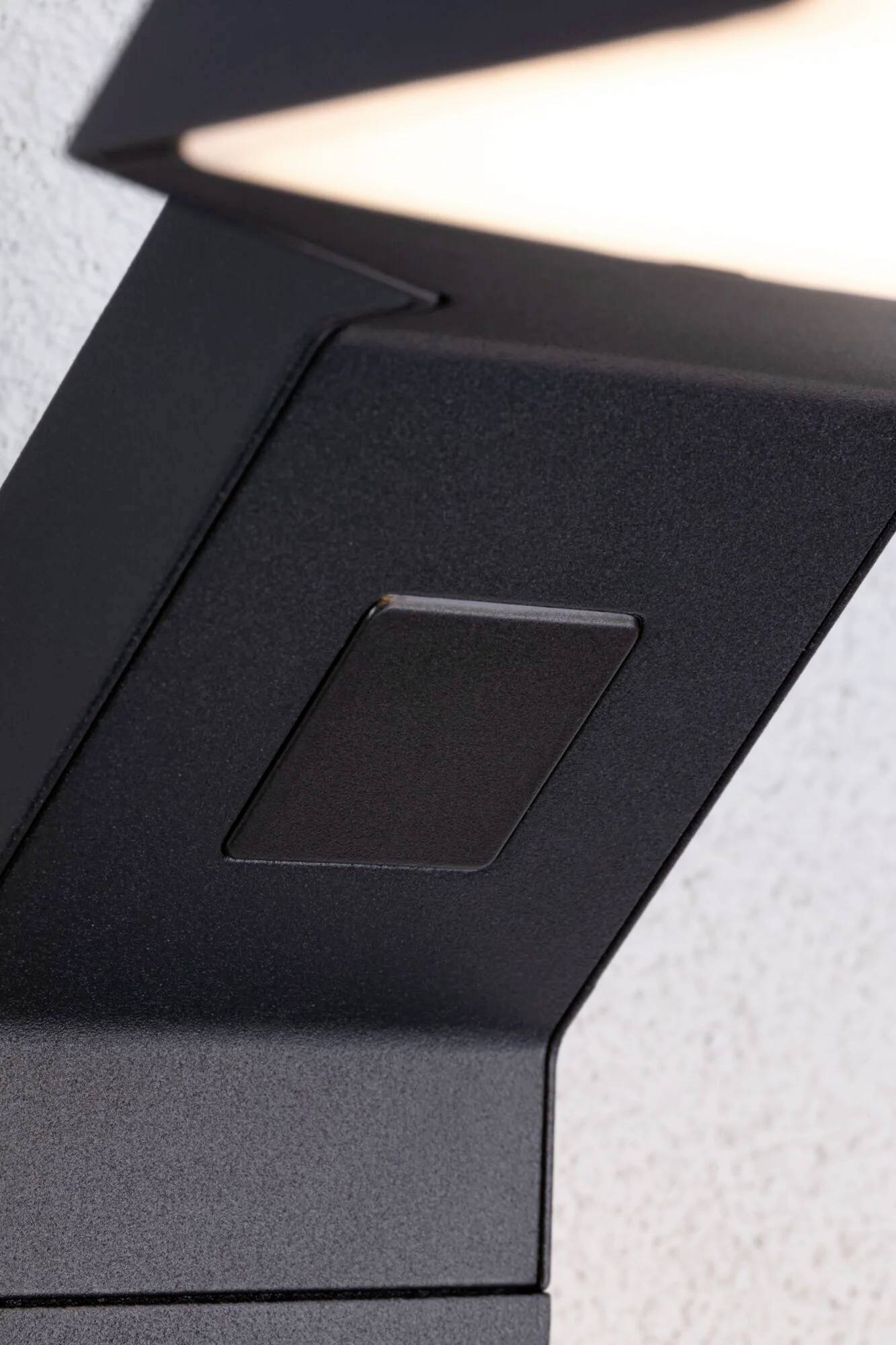 Close-up van een moderne, donkergrijze buitenlamp aan een muur, met een rechthoekige sensor onder de verlichtingskop.