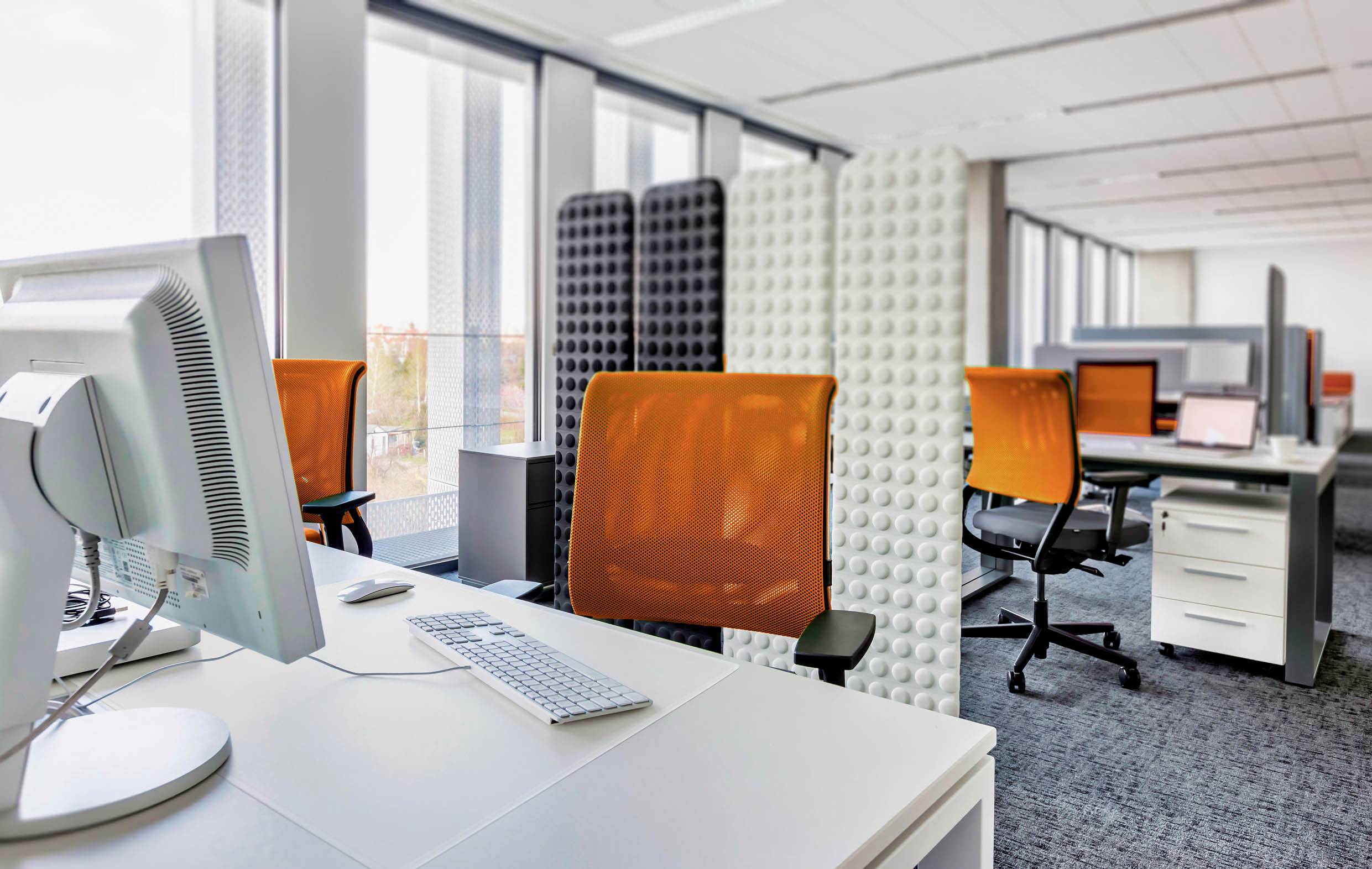 Een modern kantoor met witte tafels, oranje bureaustoelen en scheidingswanden. Daglicht valt binnen door grote ramen.