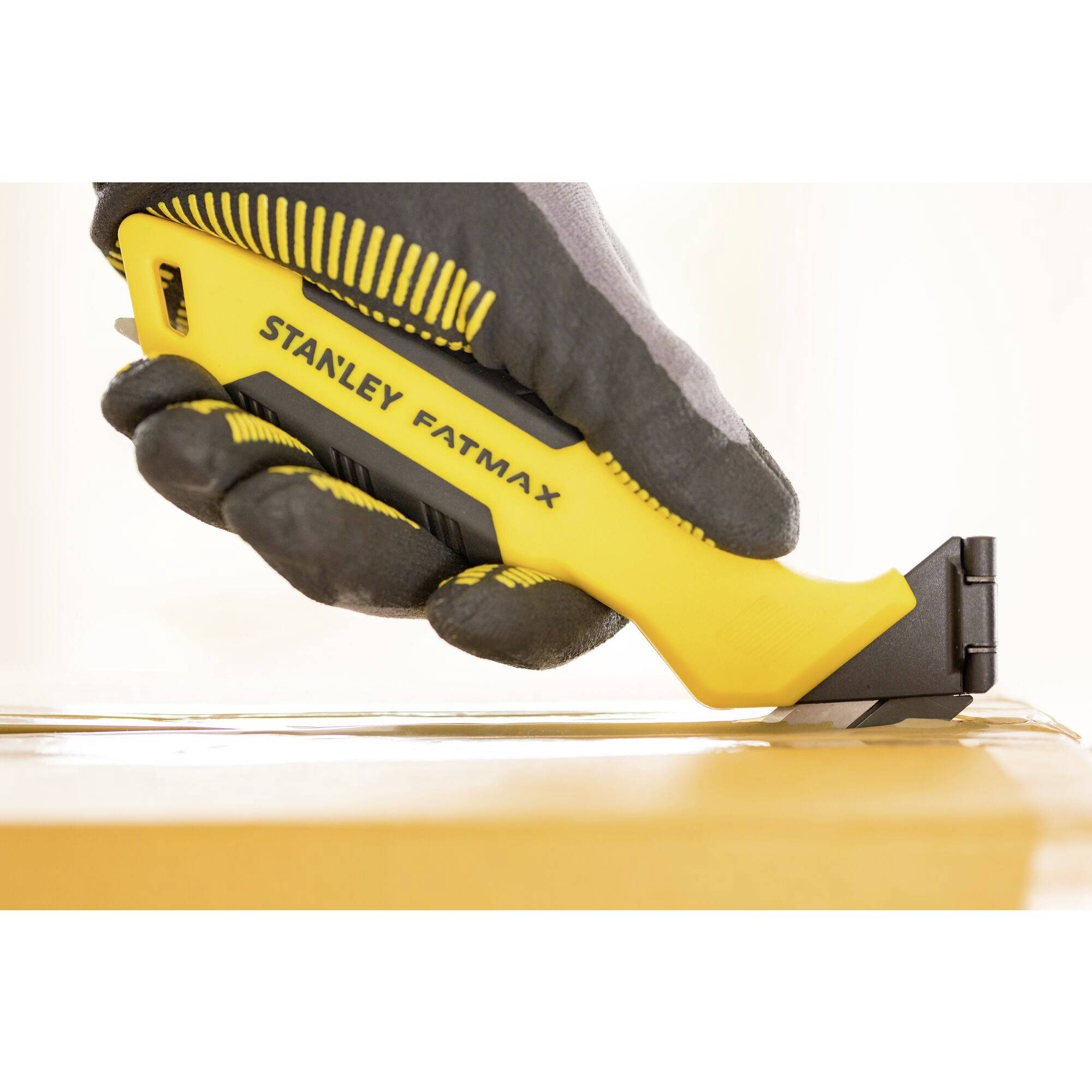 Een gele Stanley Fatmax mes met zwarte handgreep wordt gebruikt door een gehandschoende hand om een kartonnen doos te openen.
