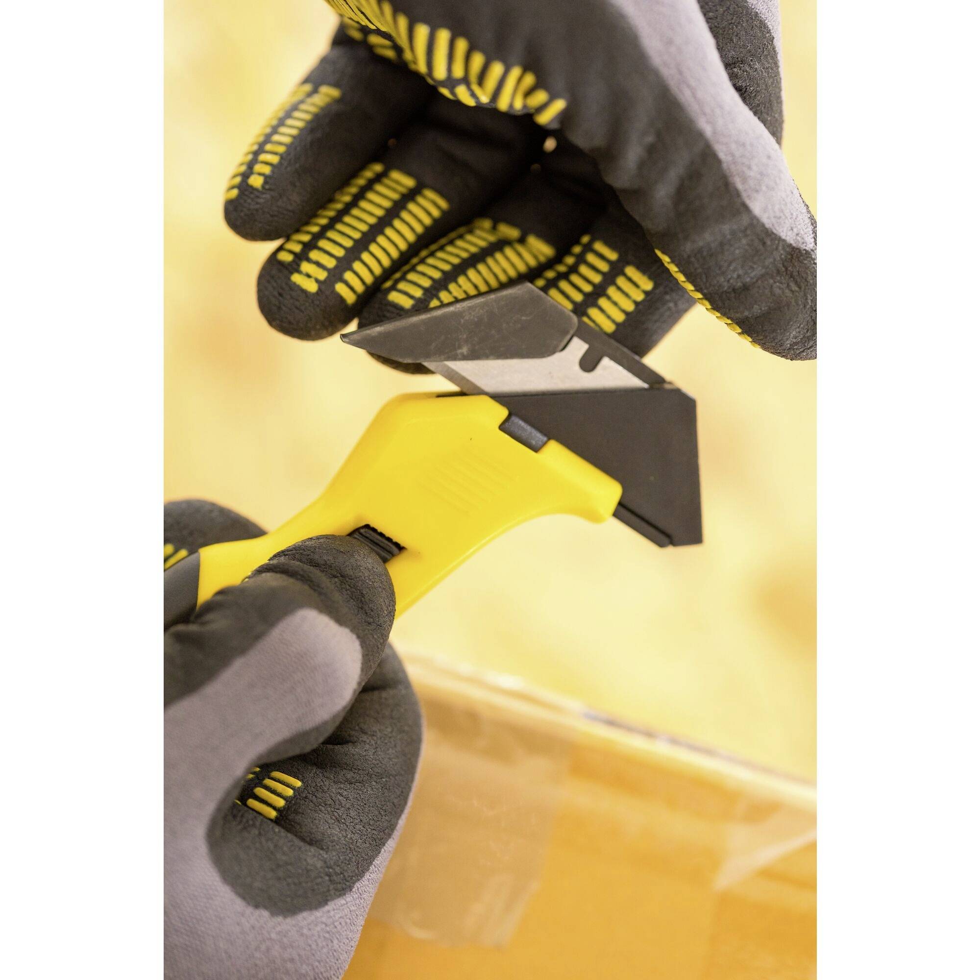 Twee gehandschoende handen snijden met een gele kartonsnijder een kartonnen doos. Veiligheidsmotief op de handschoenen.