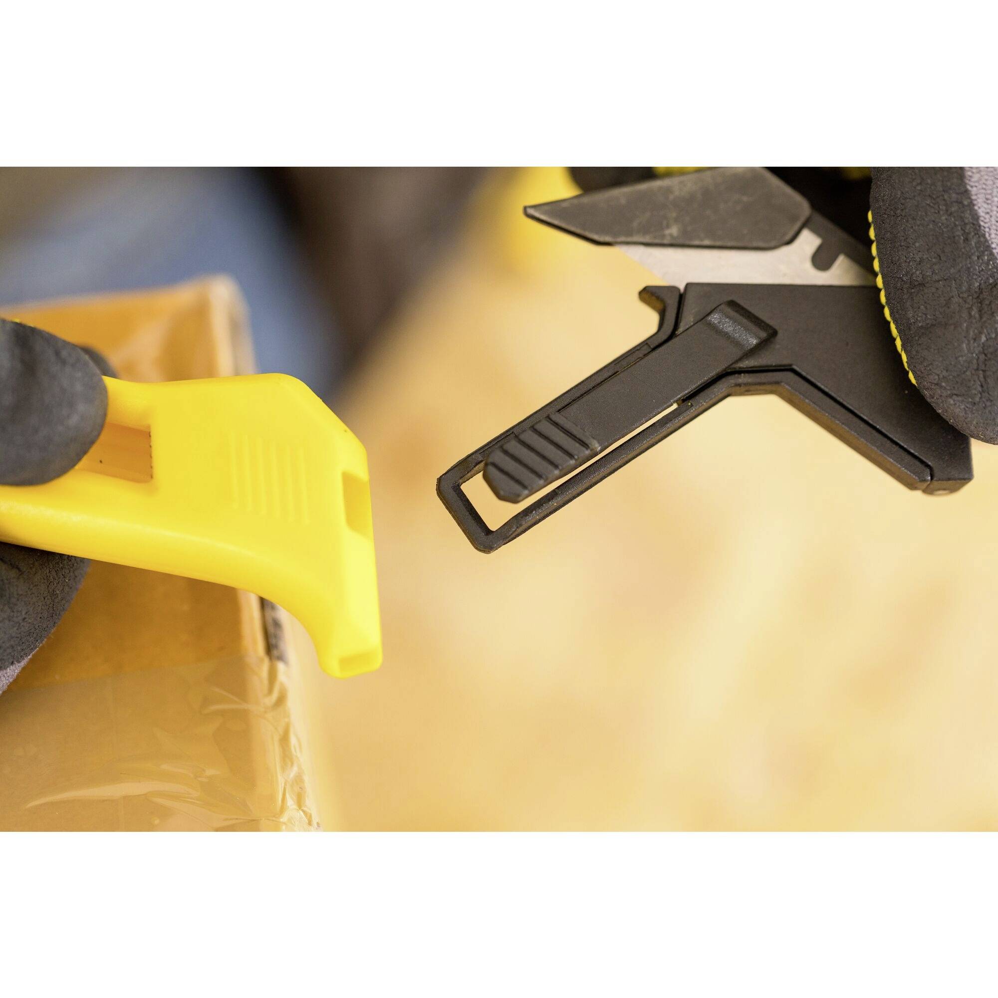 Twee handen met handschoenen houden een geel gereedschap en een tang vast, terwijl ze zich voorbereiden om een pakket te openen.