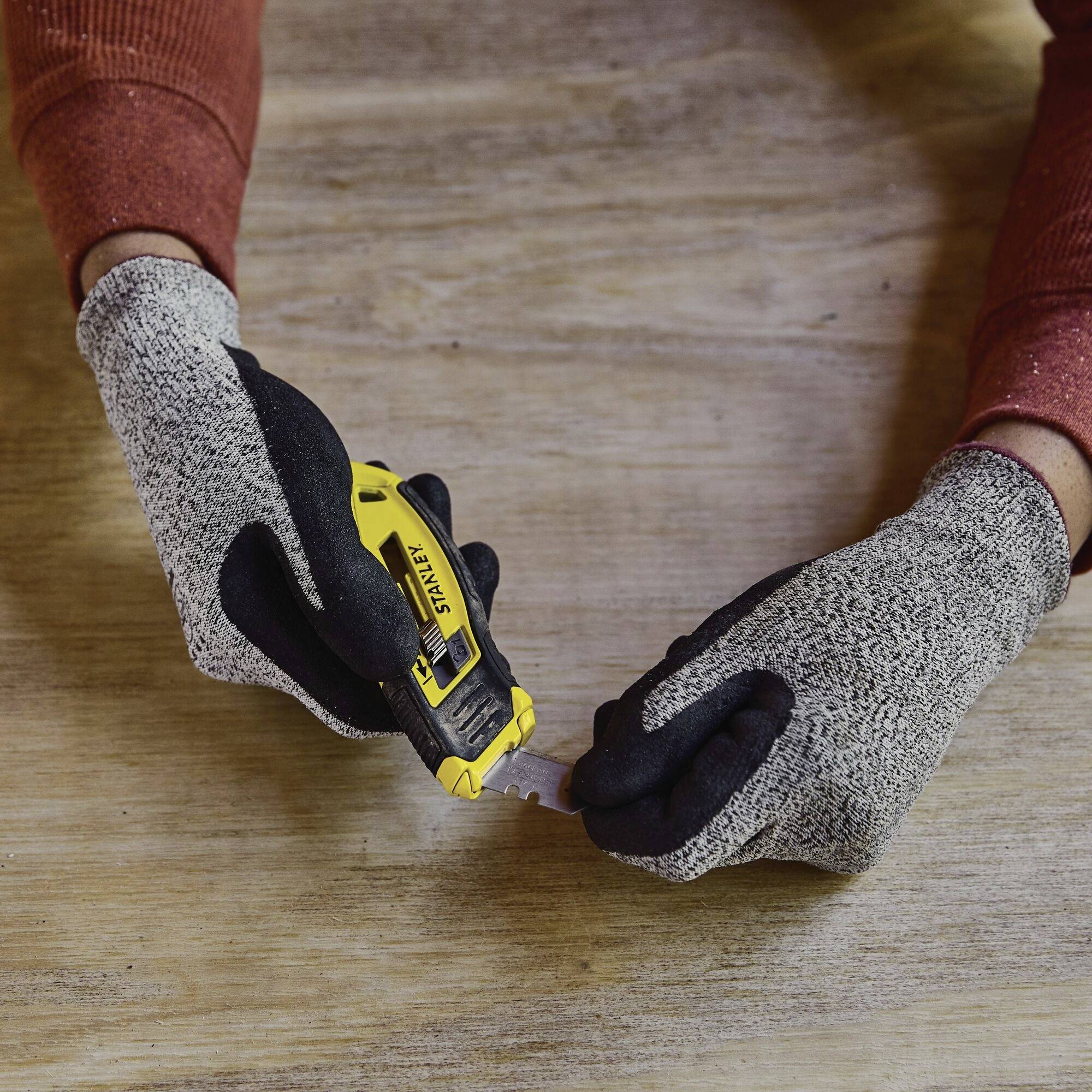 Een persoon met grijze handschoenen gebruikt een geel tapijtmes op een houten tafel.