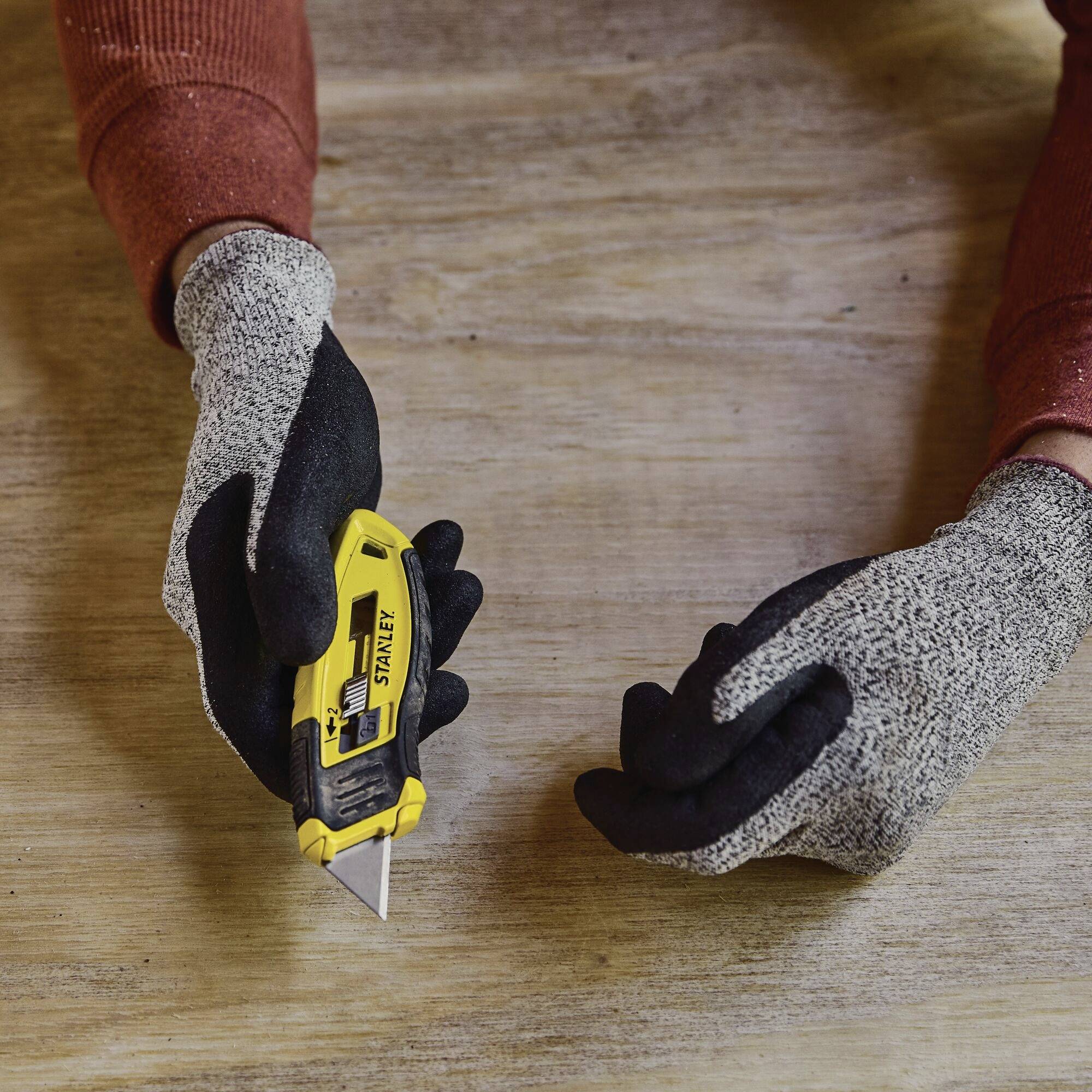 Een persoon met grijze werkhandschoenen gebruikt een geel stanleymes om een voorwerp op een houten tafel door te snijden.