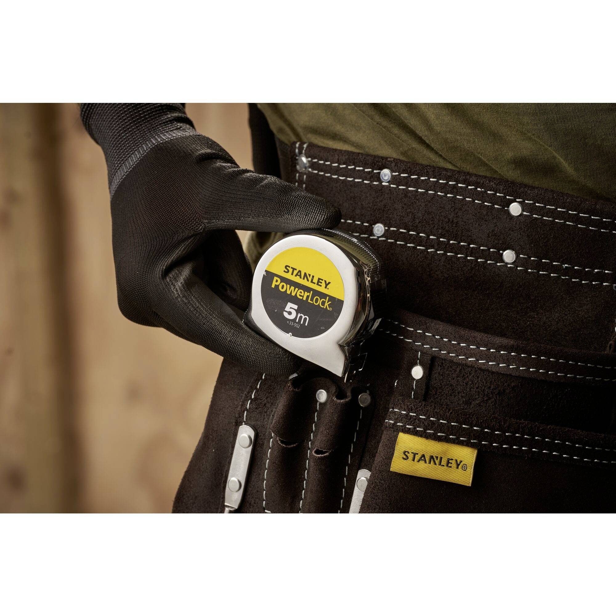 Een persoon houdt een meetlint vast met het opschrift 'STANLEY PowerLock 5m'. Hij/zij draagt handschoenen en heeft een gereedschapsriem om.