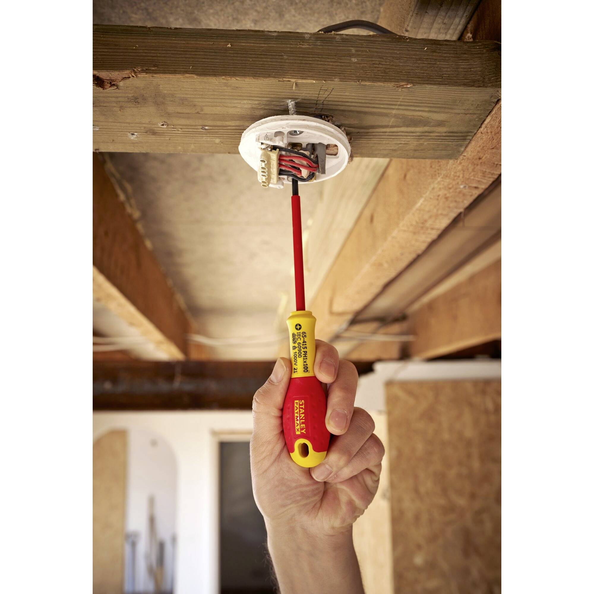 Een hand monteert een rond elektrisch stopcontact aan een houten plafond met een schroevendraaier. Draden steken uit de doos.