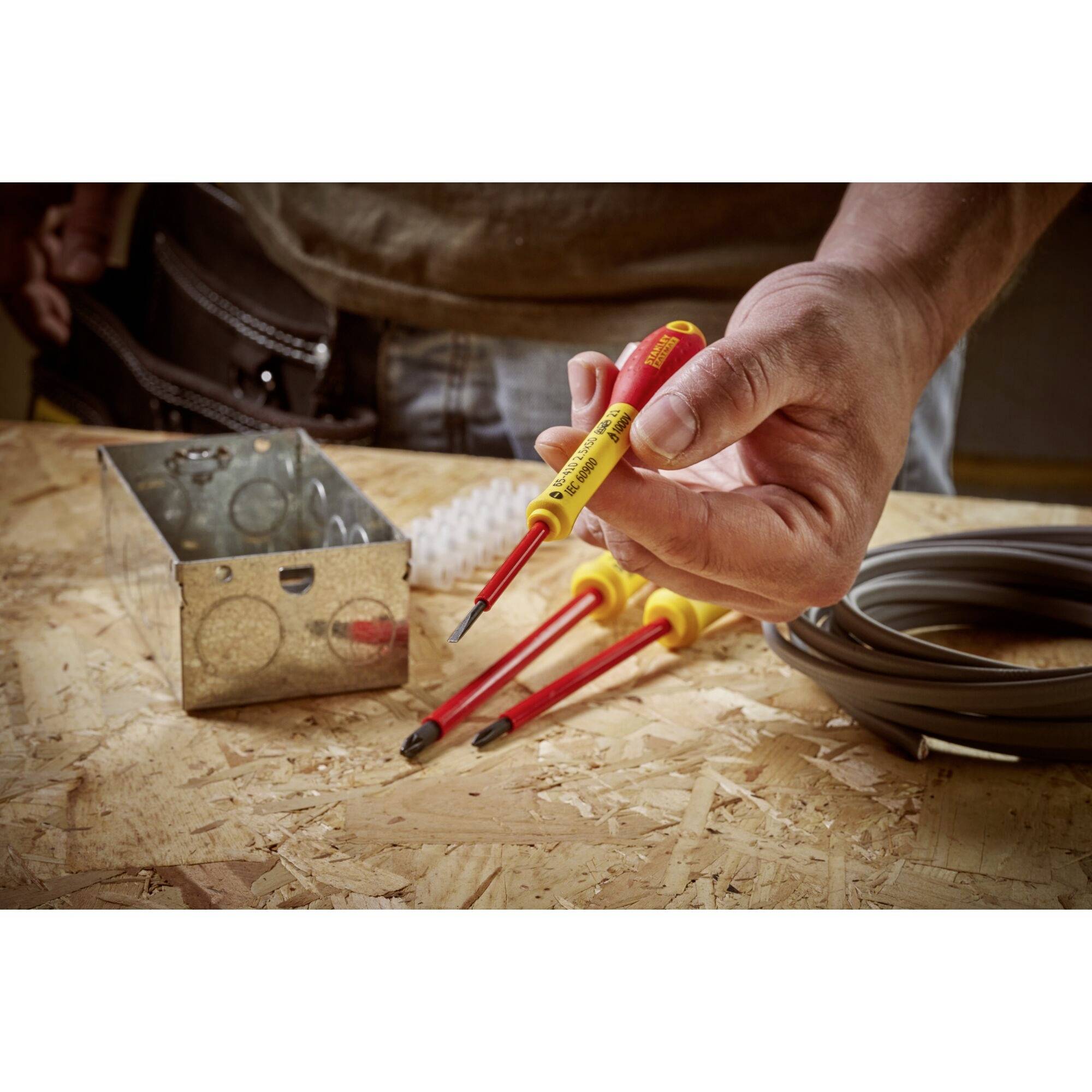 Een persoon houdt een geïsoleerde schroevendraaier. Op tafel liggen nog meer schroevendraaiers, een kabelspoel en een metalen doos.