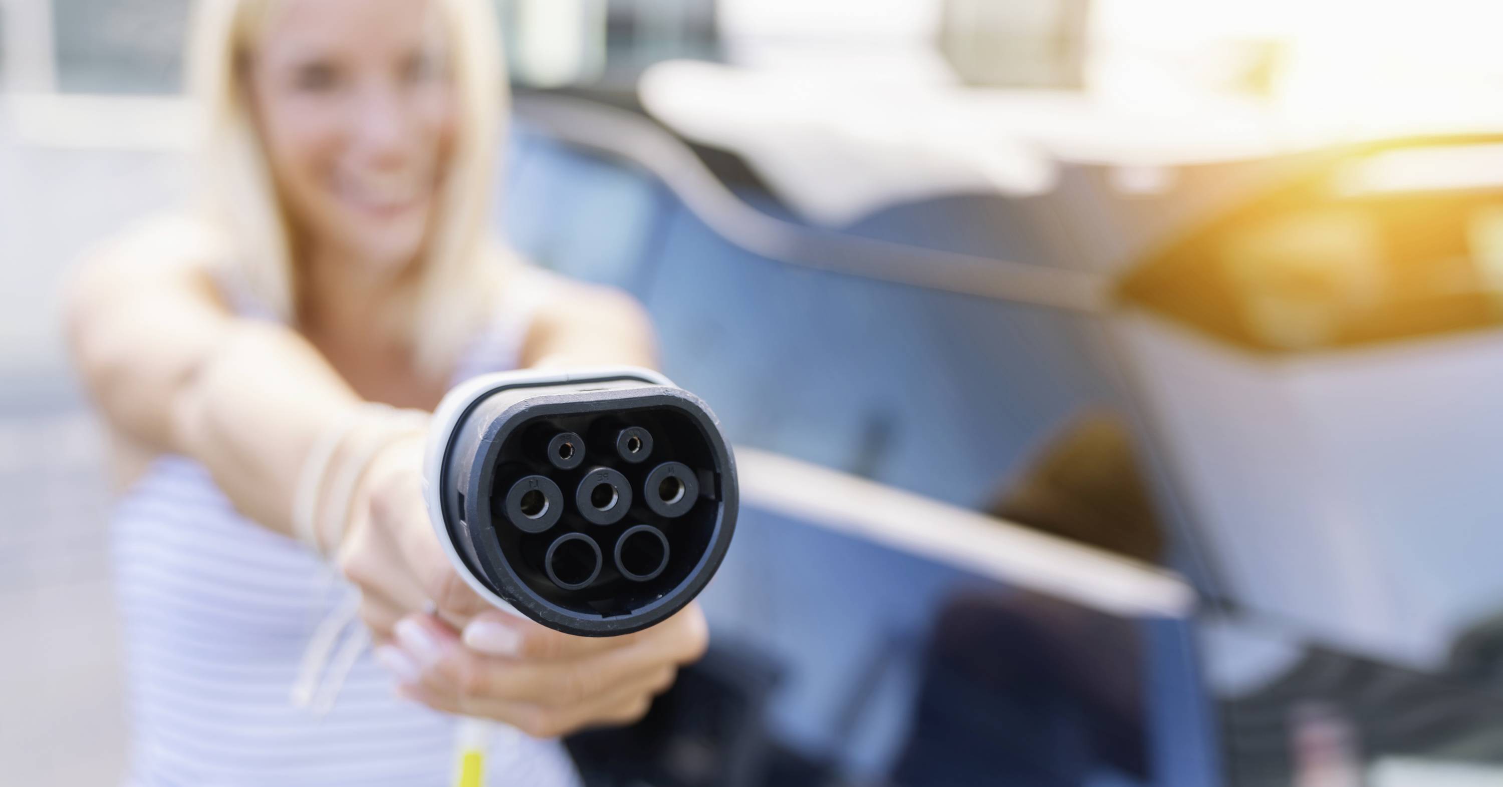 Een vrouw houdt een laadstekker voor elektrische auto's in de hand, gericht naar de toeschouwer, met op de achtergrond een auto.
