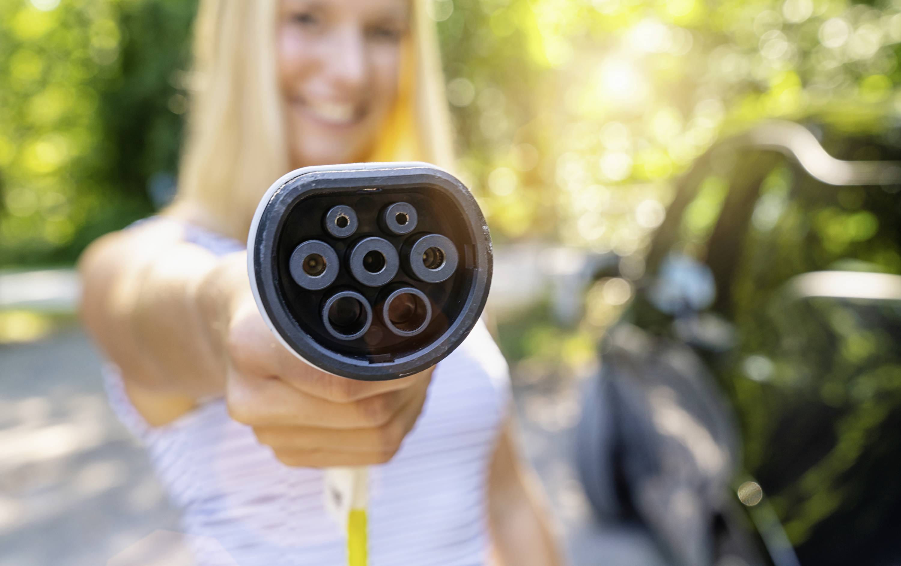 Een persoon houdt een oplaadkabel voor elektrische auto's voor de camera. Op de achtergrond wazig een auto en bomen in zonlicht.