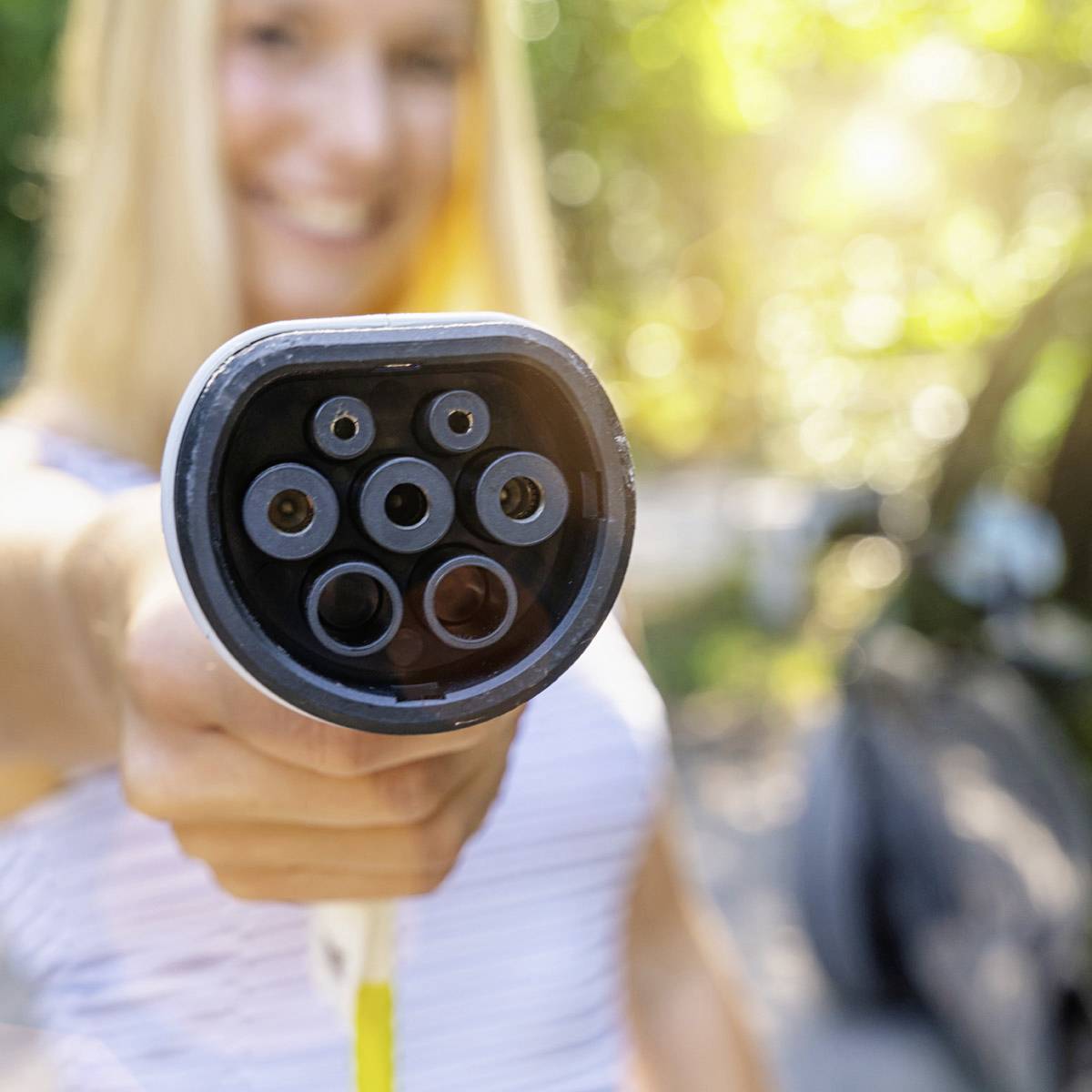 Een persoon houdt een laadstekker voor een elektrisch voertuig in de hand. Op de achtergrond wazig een elektrische auto zichtbaar in een zonnige omgeving.