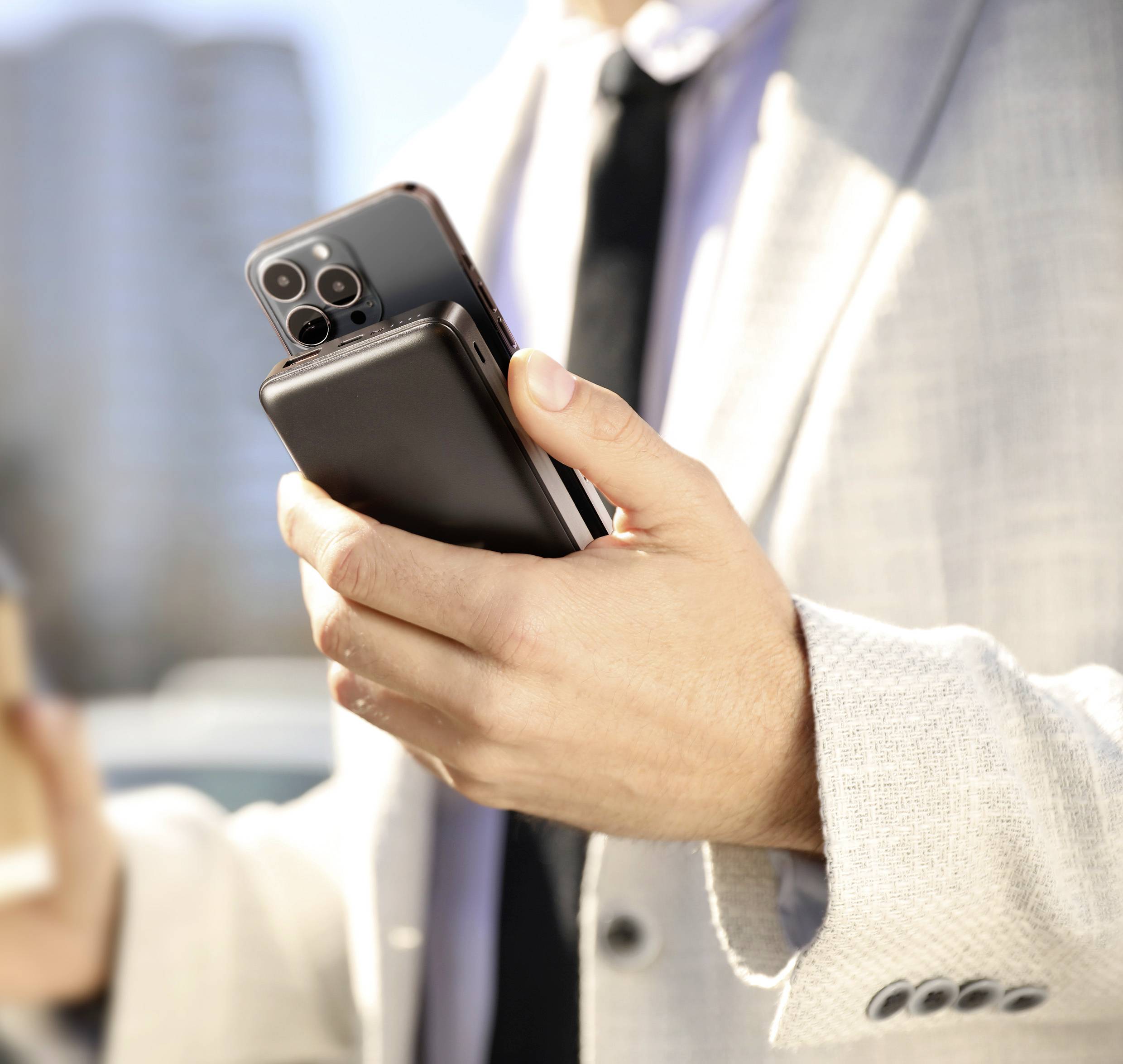 Een persoon in een pak houdt twee smartphones en een koffiekopje vast. Op de achtergrond zijn wazig gebouwen zichtbaar.