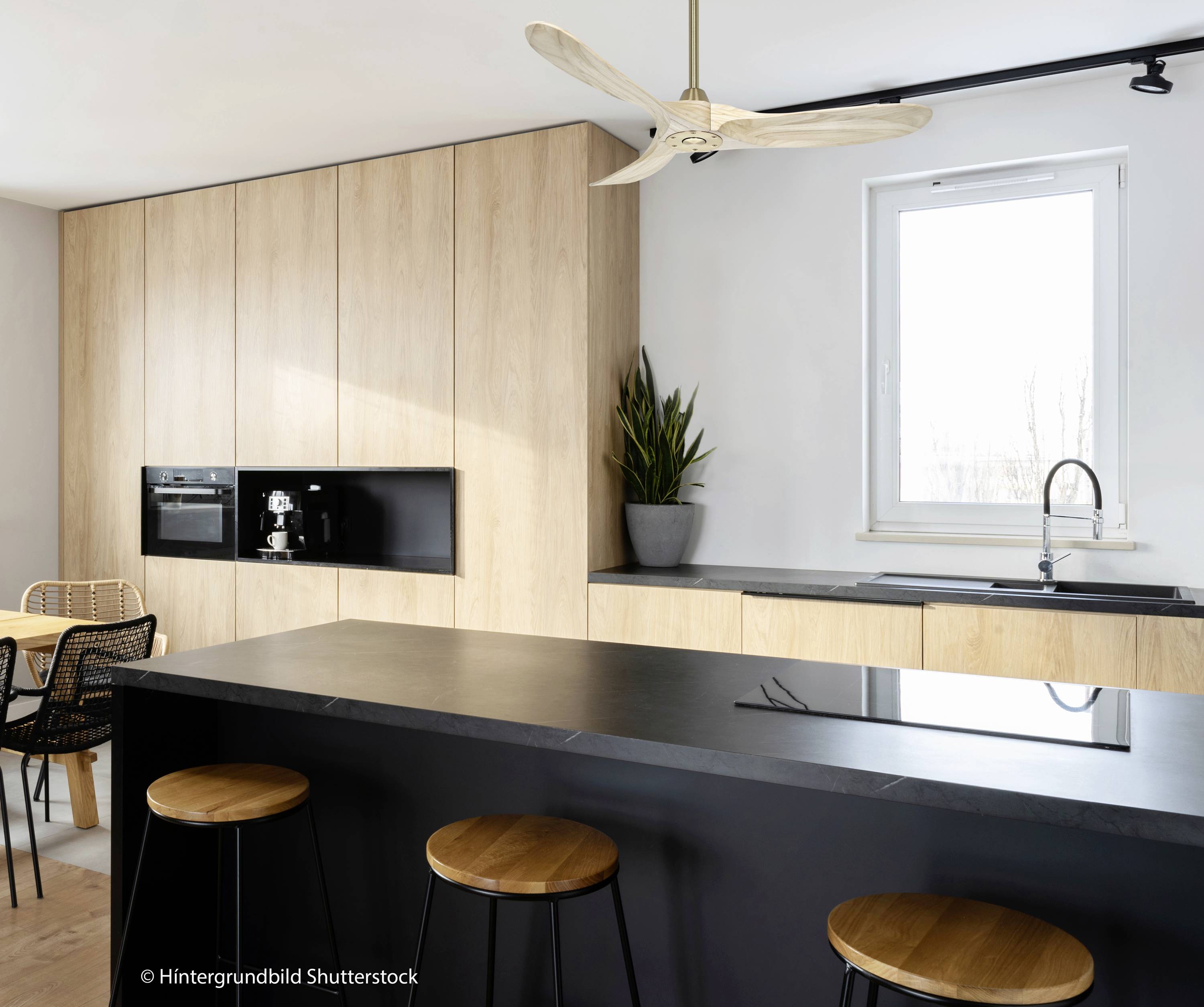 Moderne keuken met zwart werkblad en houten decor. Zwarte barkrukken voor het keukeneiland, planten en ramen op de achtergrond.