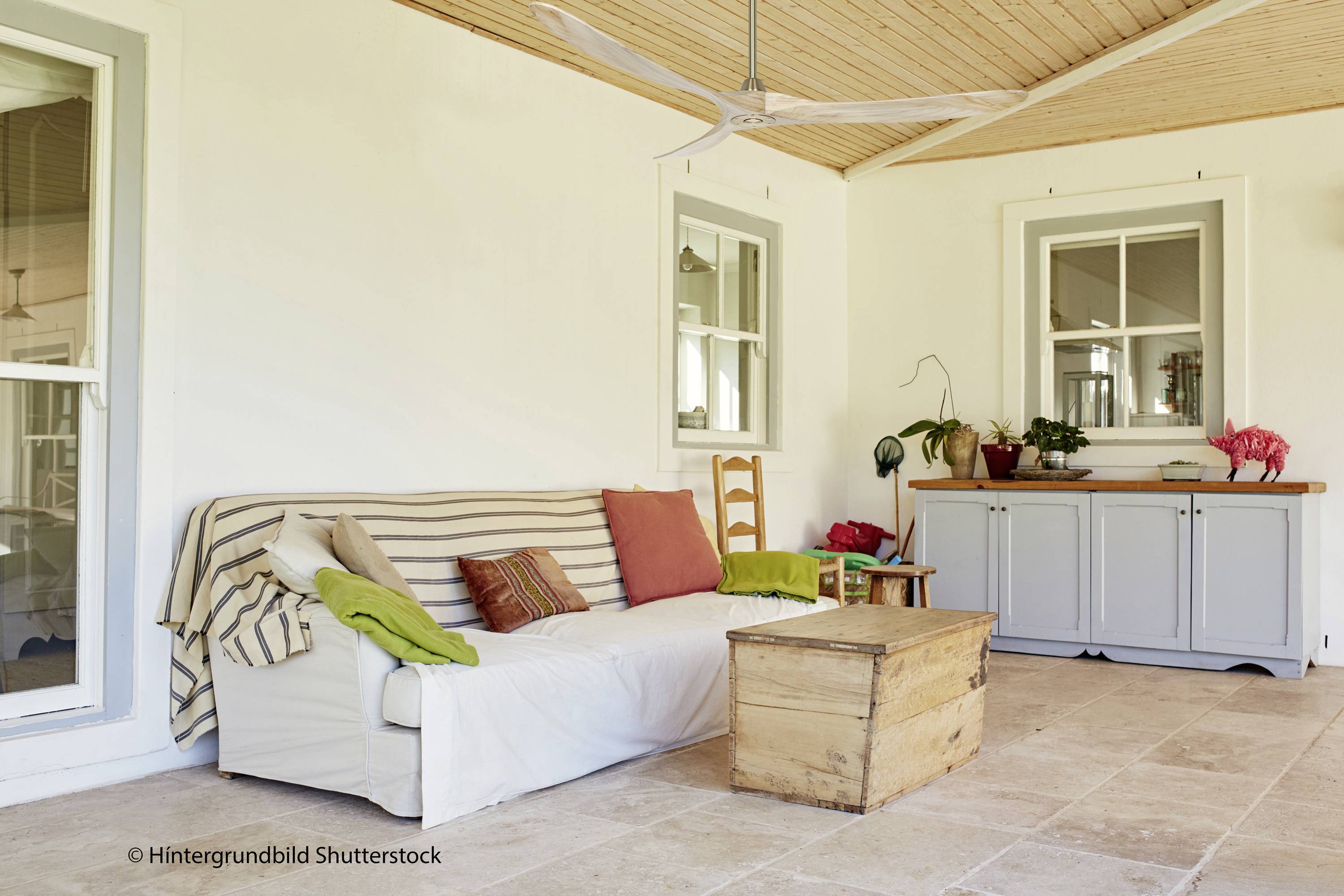Een gezellige binnenplaats met een witte bank, gekleurde kussens, een houten tafel en planten op een dressoir voor een witte muur.