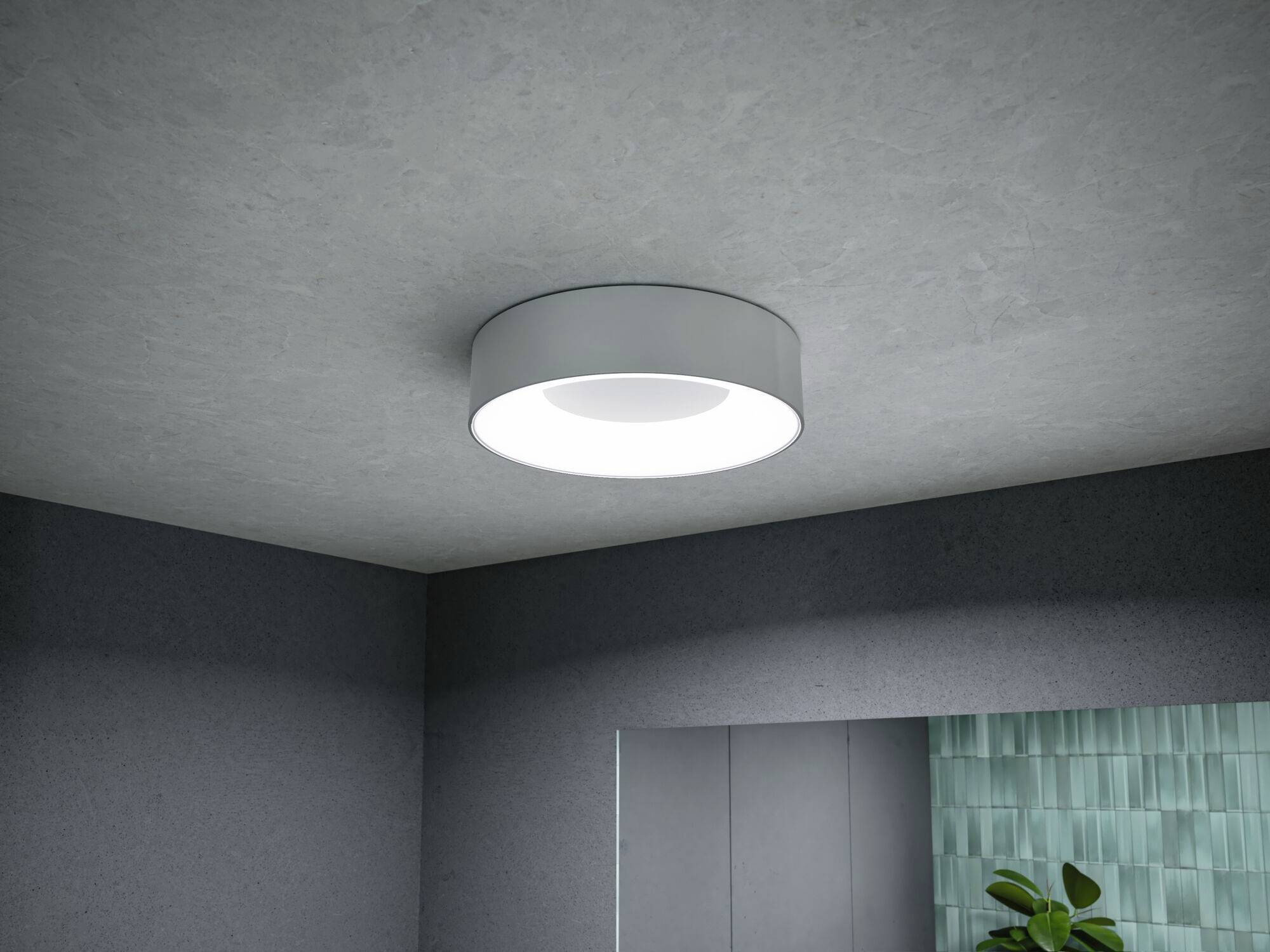 Een ronde, moderne plafondlamp met helder licht schijnt aan het grijze plafond van een ruimte met een plant en een betegelde muur.