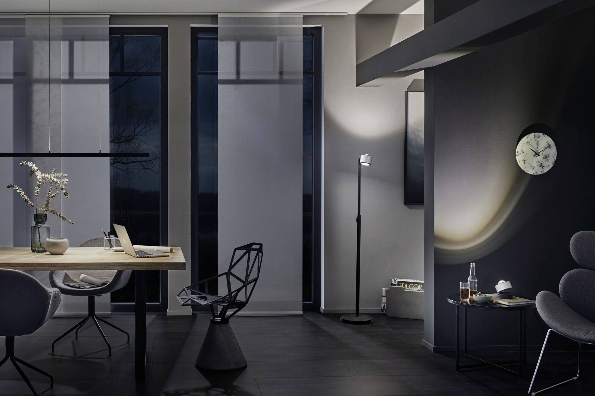 Modern woonkamer met donkere vloer, een houten tafel, moderne stoelen, staande lamp, klok aan de muur en een open laptop op tafel.