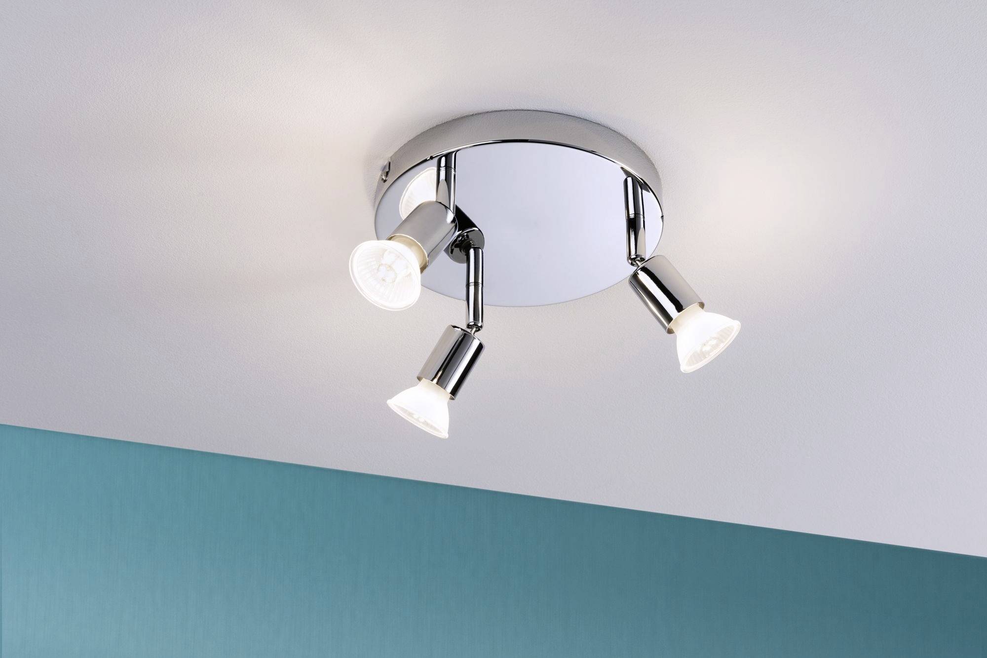 Drievoudige plafondlamp met verstelbare spots in modern design, gemonteerd aan een witte plafond boven een turquoise muur.