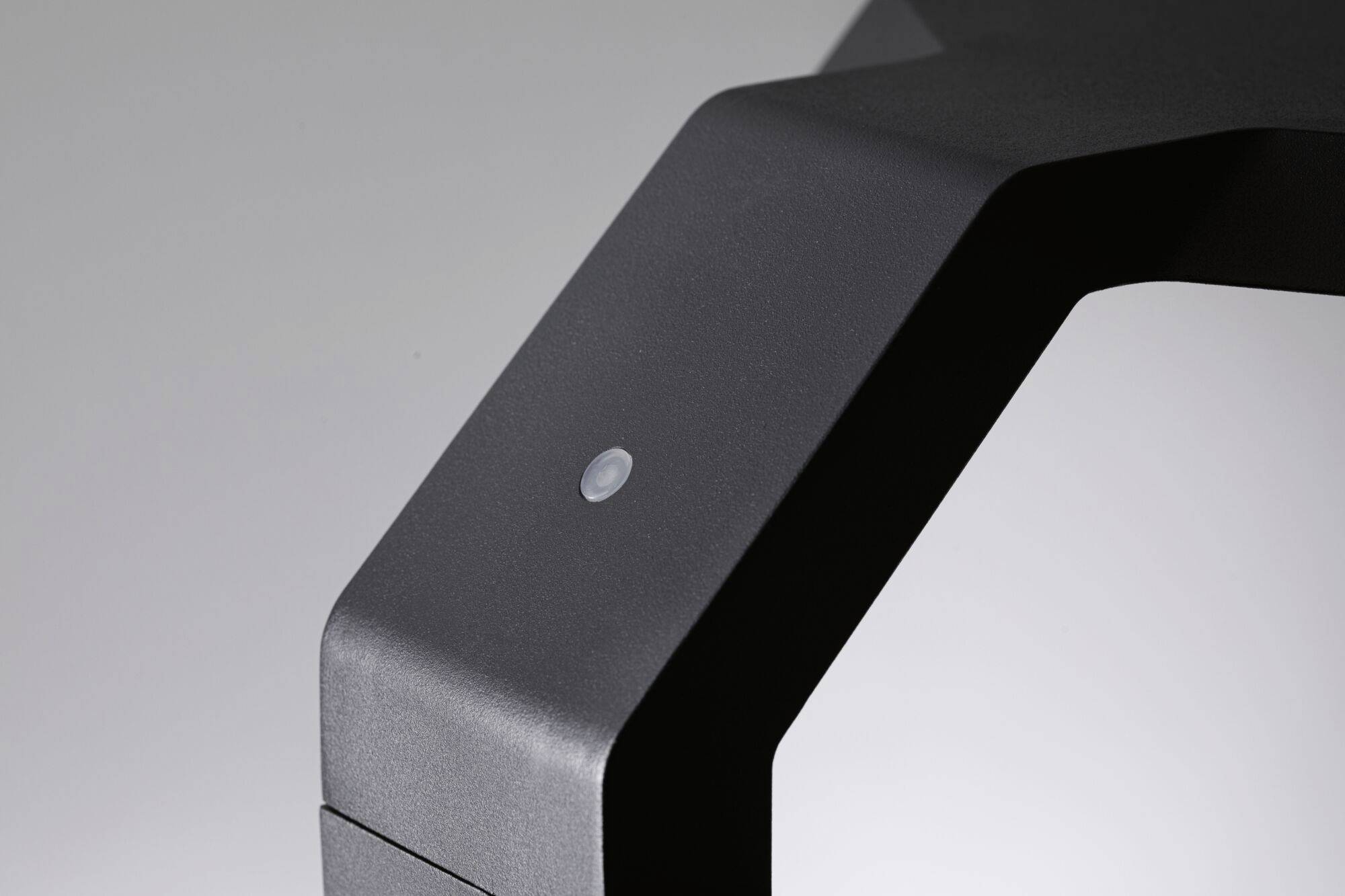 Donker, hoekig metalen onderdeel met een kleine knop in het midden. De achtergrond is grijs.